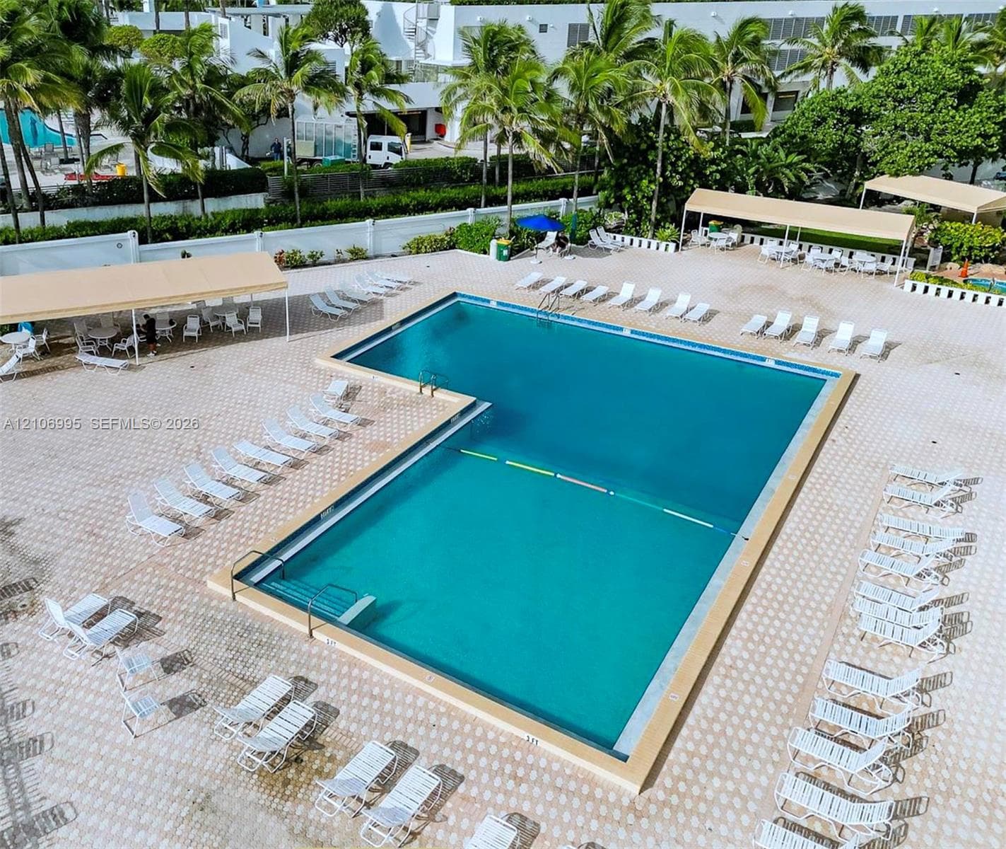 L-shaped swimming pool featuring an integrated staircase, expansive paved patio, multiple covered cabanas, and perimeter landscaping