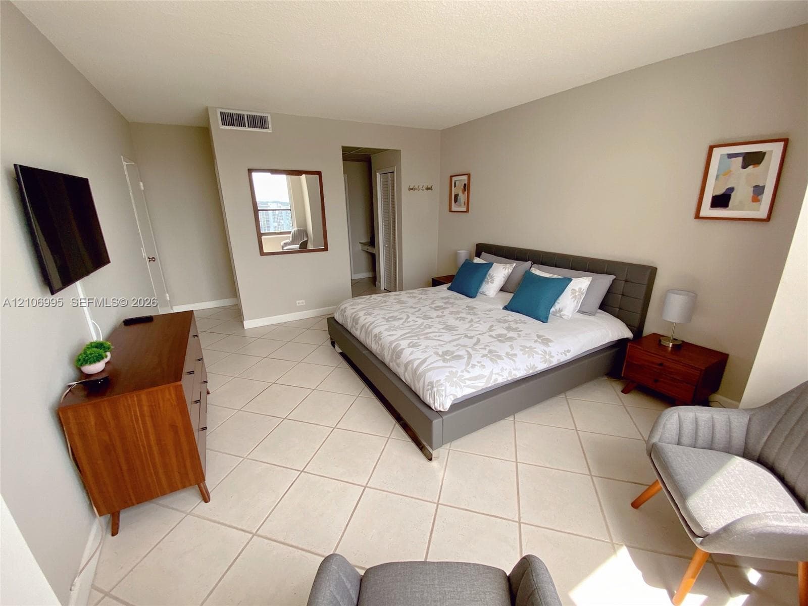 Neutral-toned interior space featuring light-colored diagonal floor tiling, wall-mounted mirror, and wall-mounted television