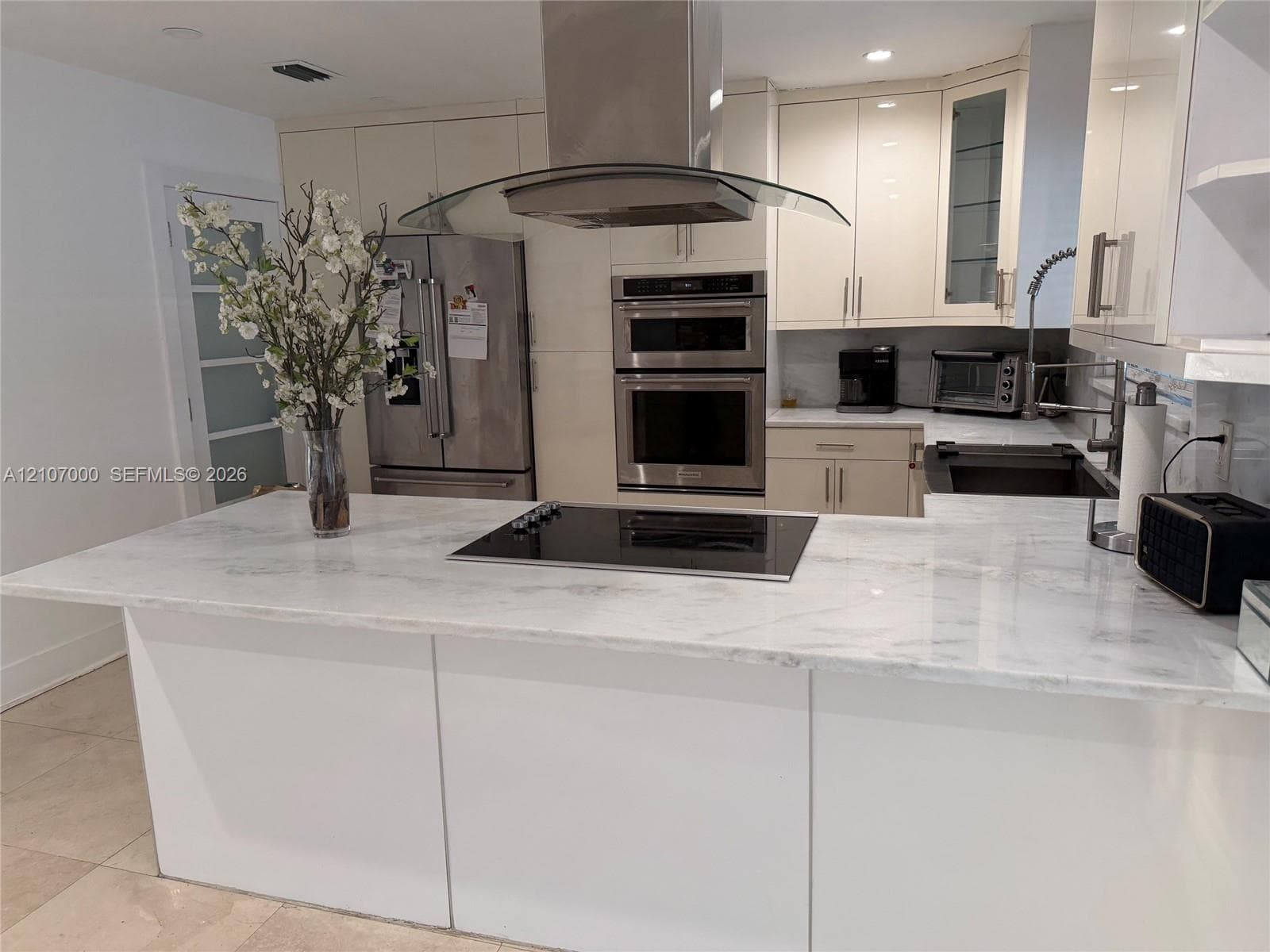 Kitchen featuring a stone-surface island with an integrated cooktop, stainless steel double wall ovens, a glass and stainless steel vent hood, and high-gloss cabinetry