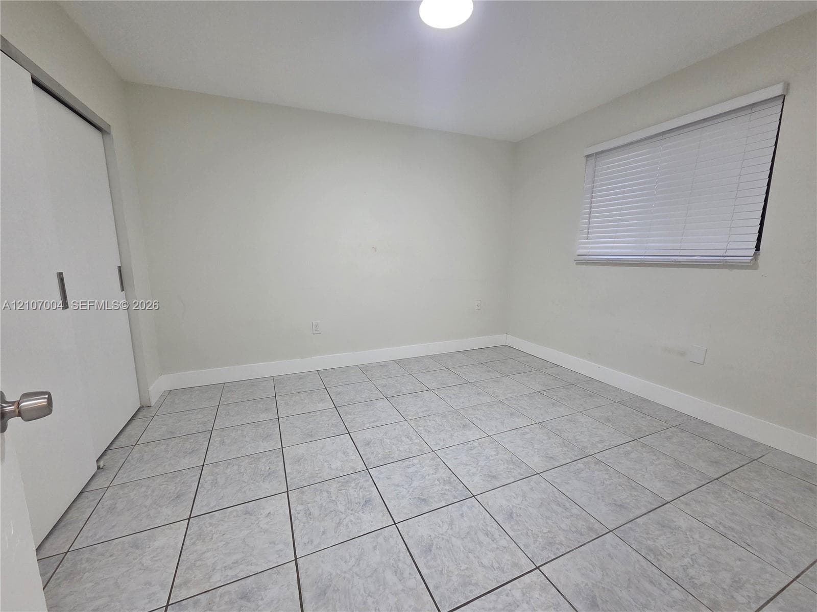 Neutral-toned room featuring large-format square tile flooring, a dual-panel closet door, and a horizontal slatted window blind