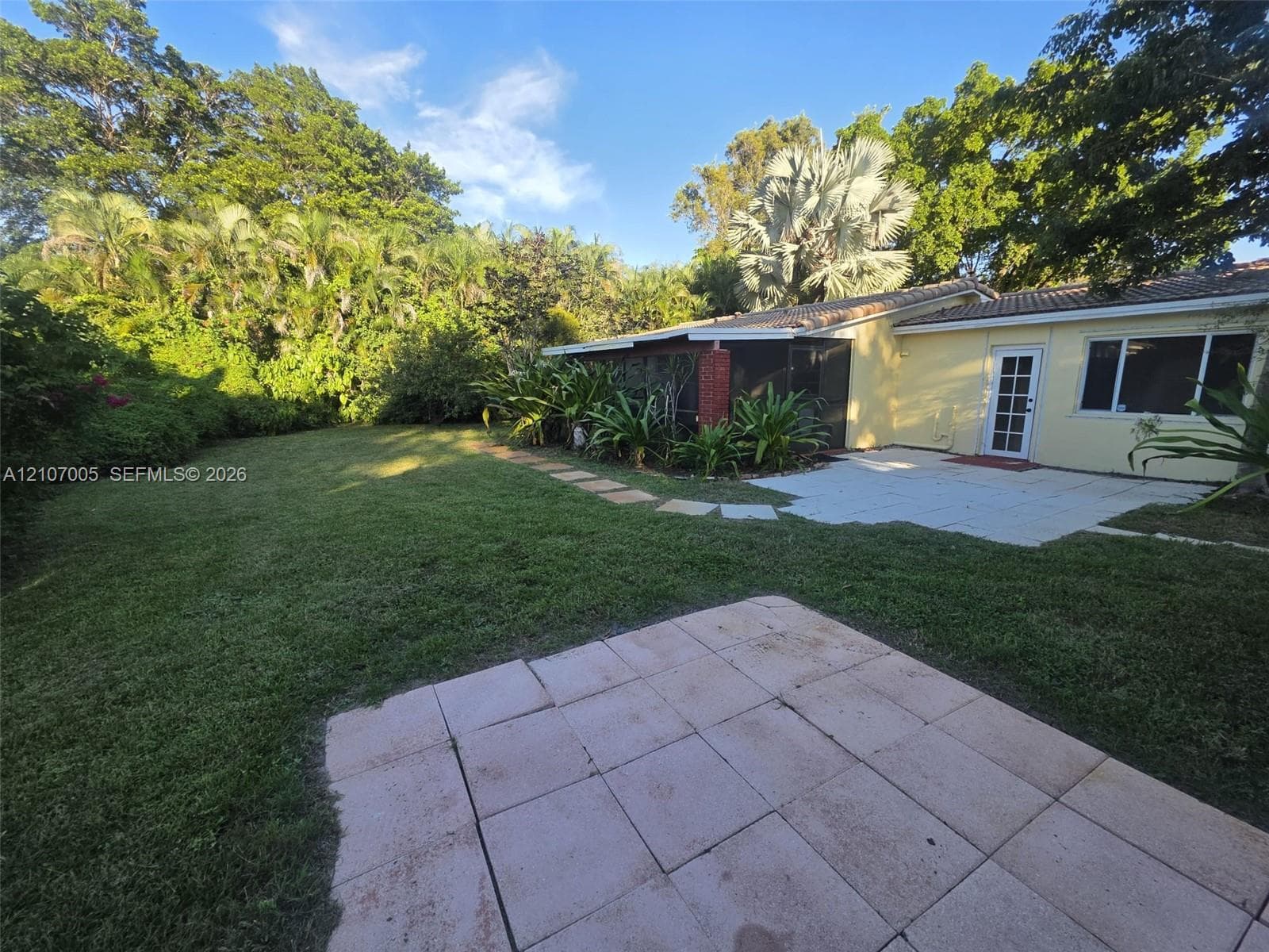 Spacious backyard featuring a grass lawn, paved stone patio, screened porch, and perimeter landscaping with mature trees and foliage