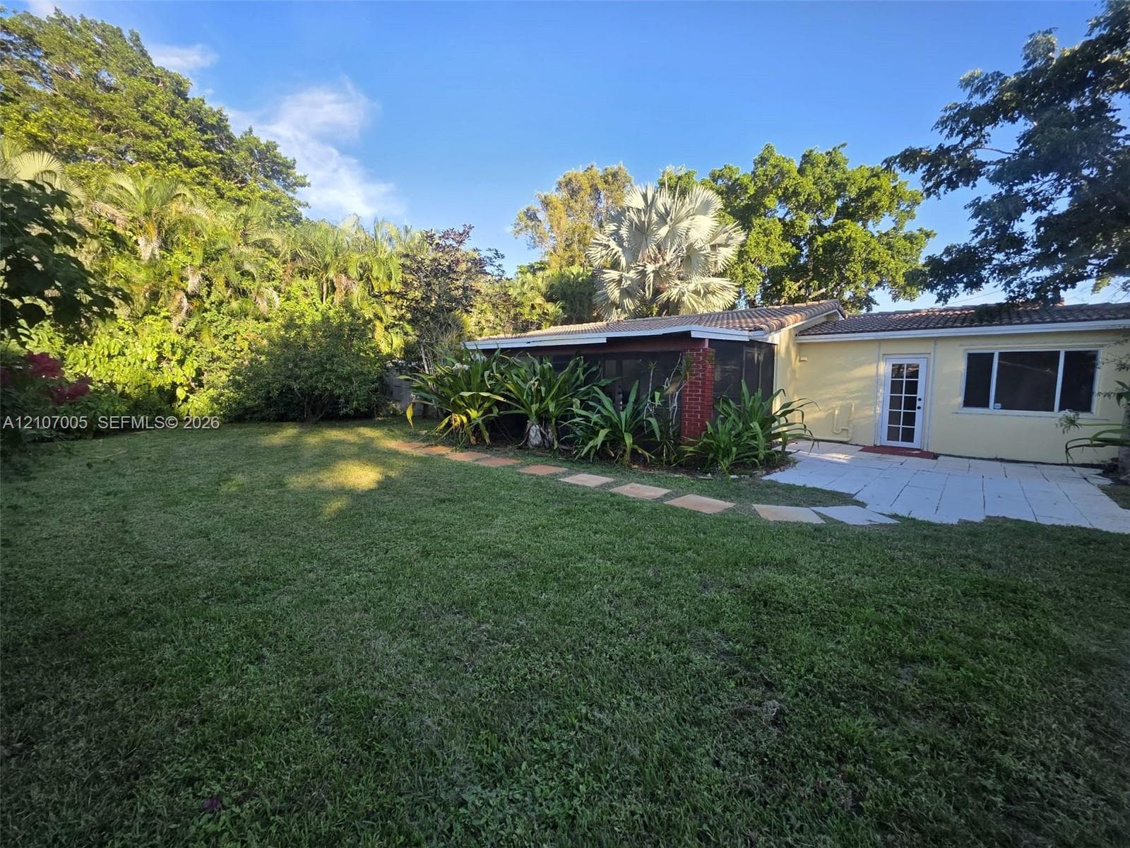 Spacious backyard featuring a manicured lawn, stone paver path, paved patio, and mature tropical landscaping