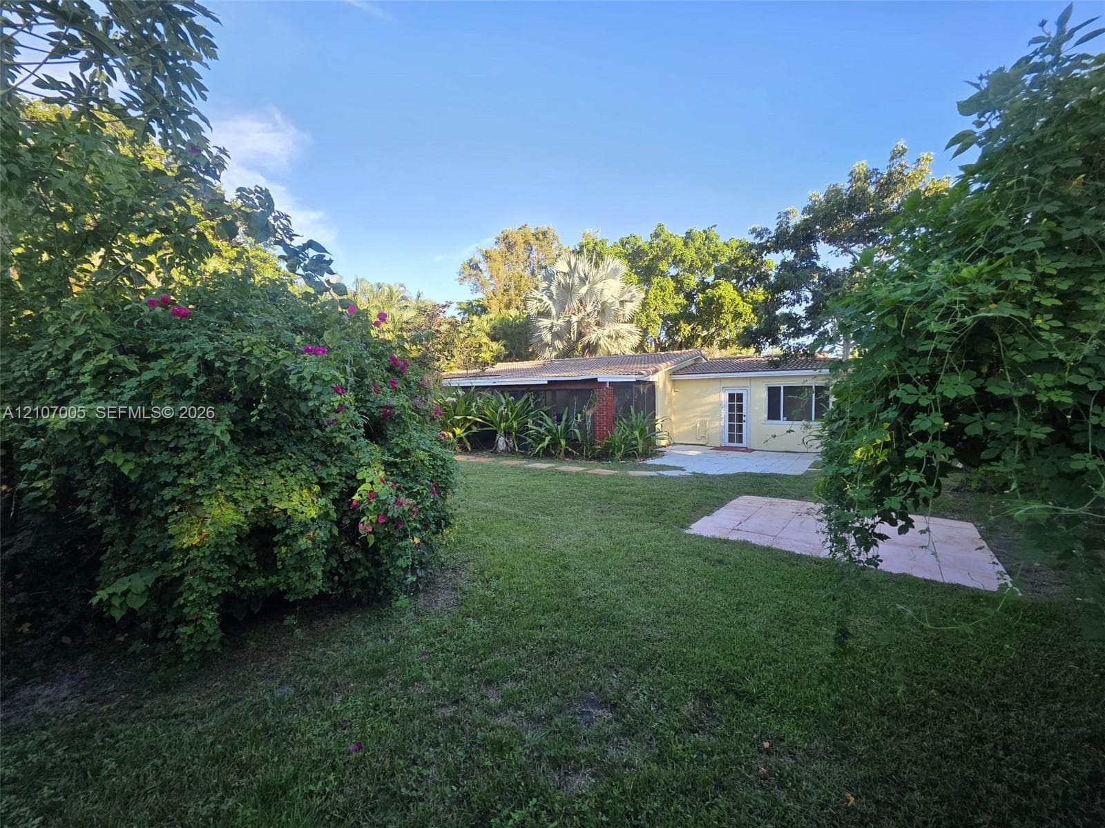 Backyard featuring a grass lawn, mature trees, flowering shrubbery, and a concrete patio