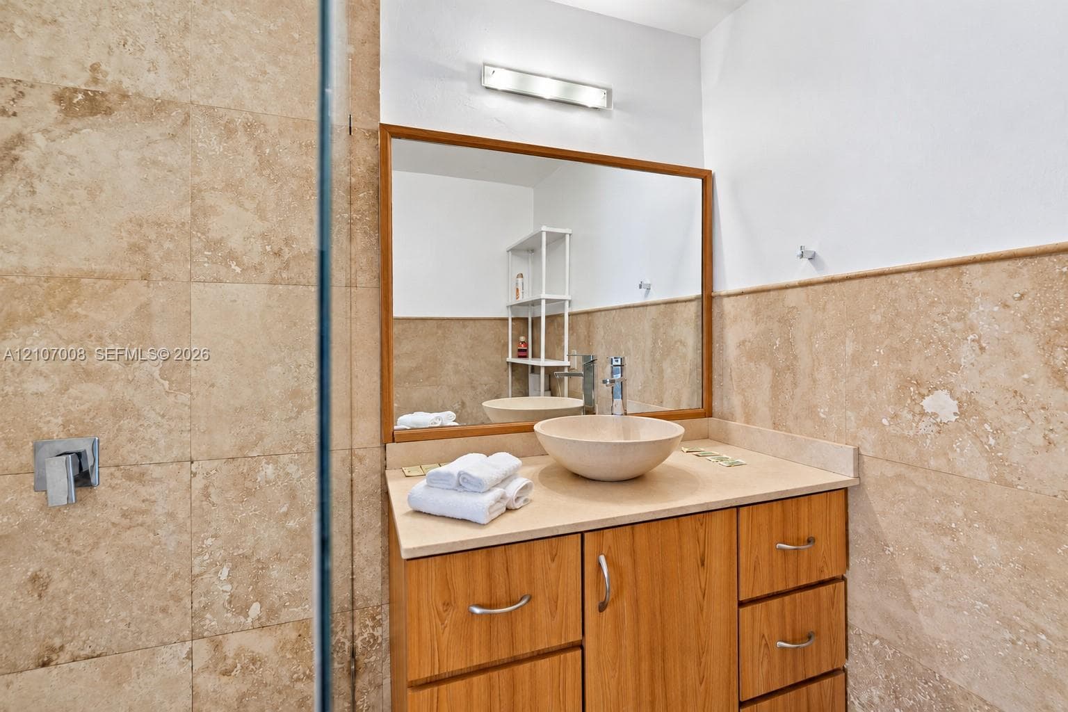 Vanity featuring wood-finish cabinetry, a stone-composite countertop, a circular vessel sink, a chrome single-handle faucet, and large-format neutral-toned tile wall cladding