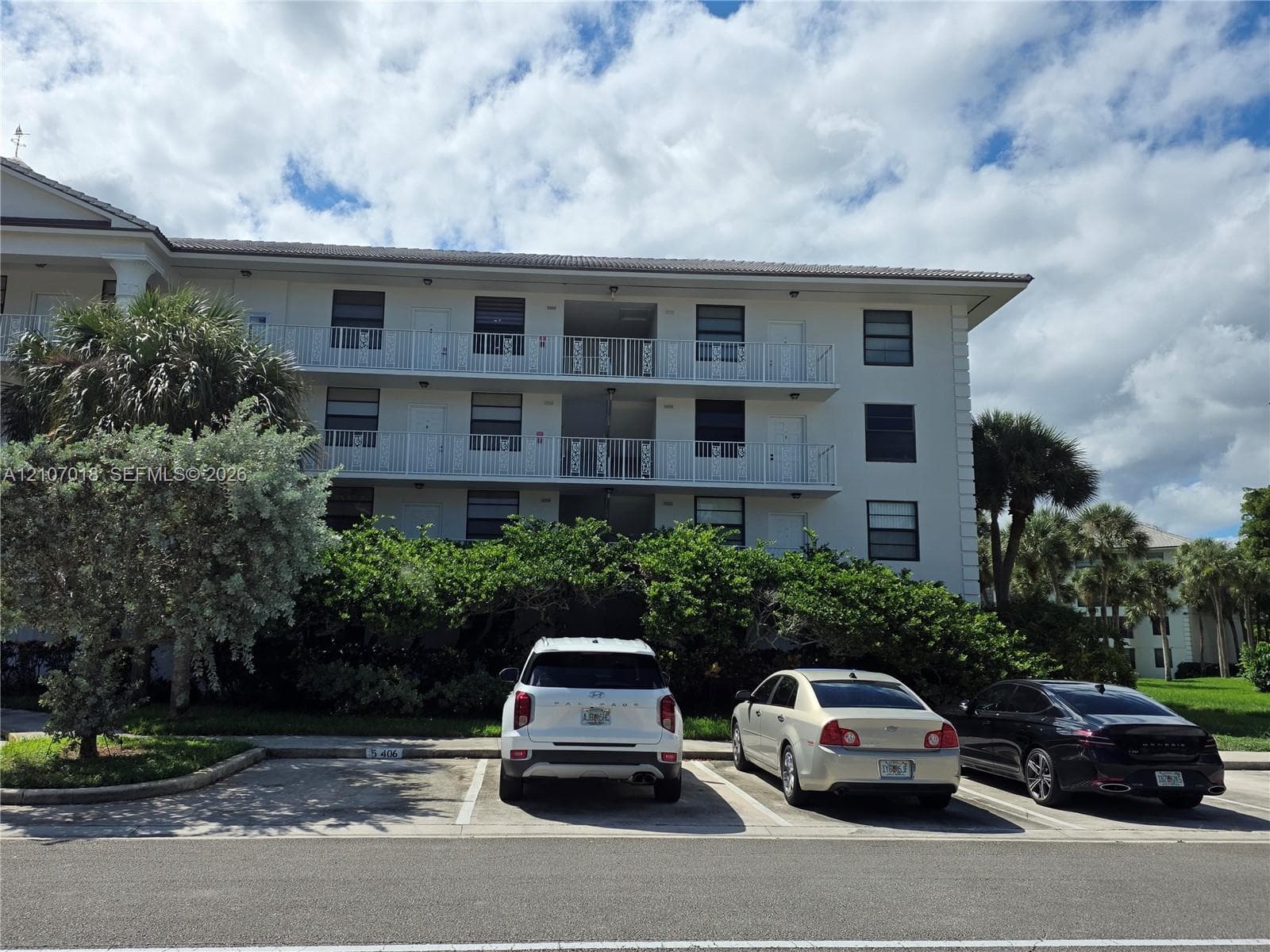Multi-story residential building exterior featuring a white stucco facade, shingle roof, integrated metal balcony railings, and assigned surface parking spaces