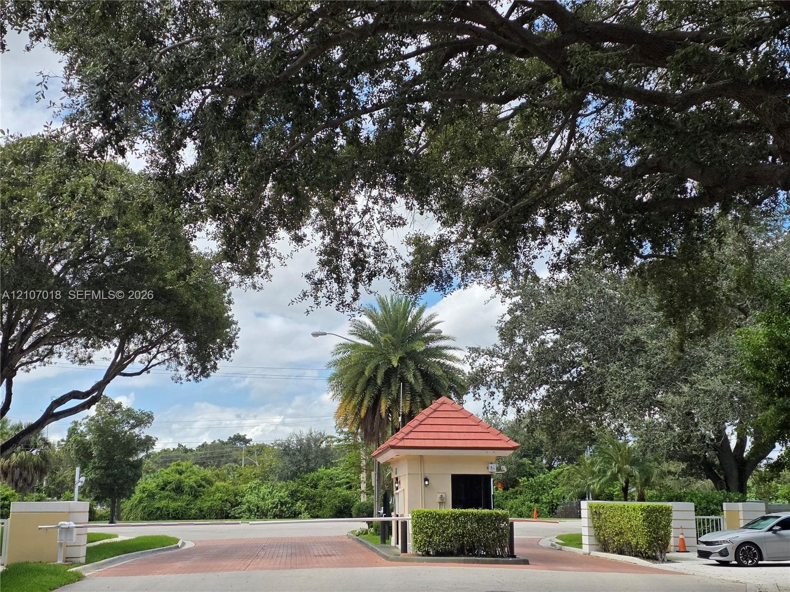 Gated entrance featuring a guardhouse with a tiled roof, paved brick thoroughfare, and mature landscape canopy