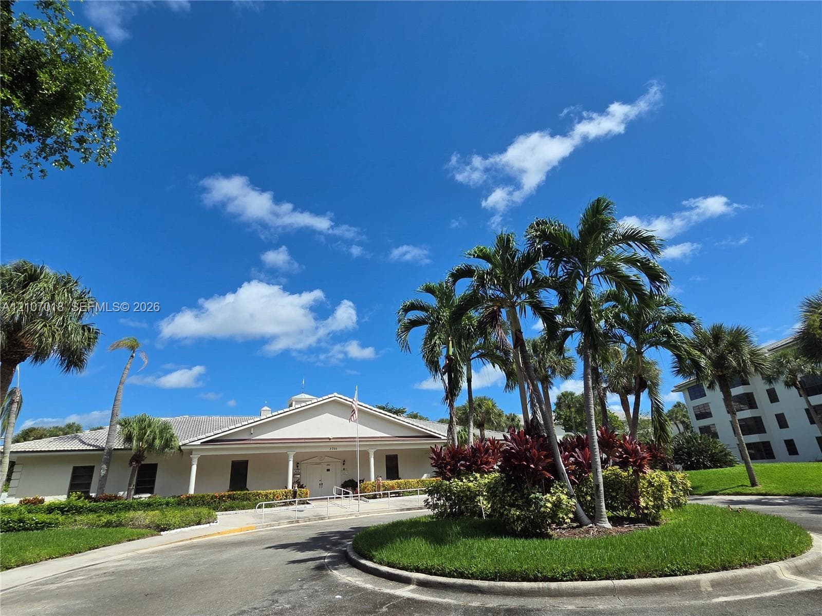 Exterior featuring a light-toned stucco façade, a multi-gabled roofline, covered entryway with support columns, and mature tropical landscaping