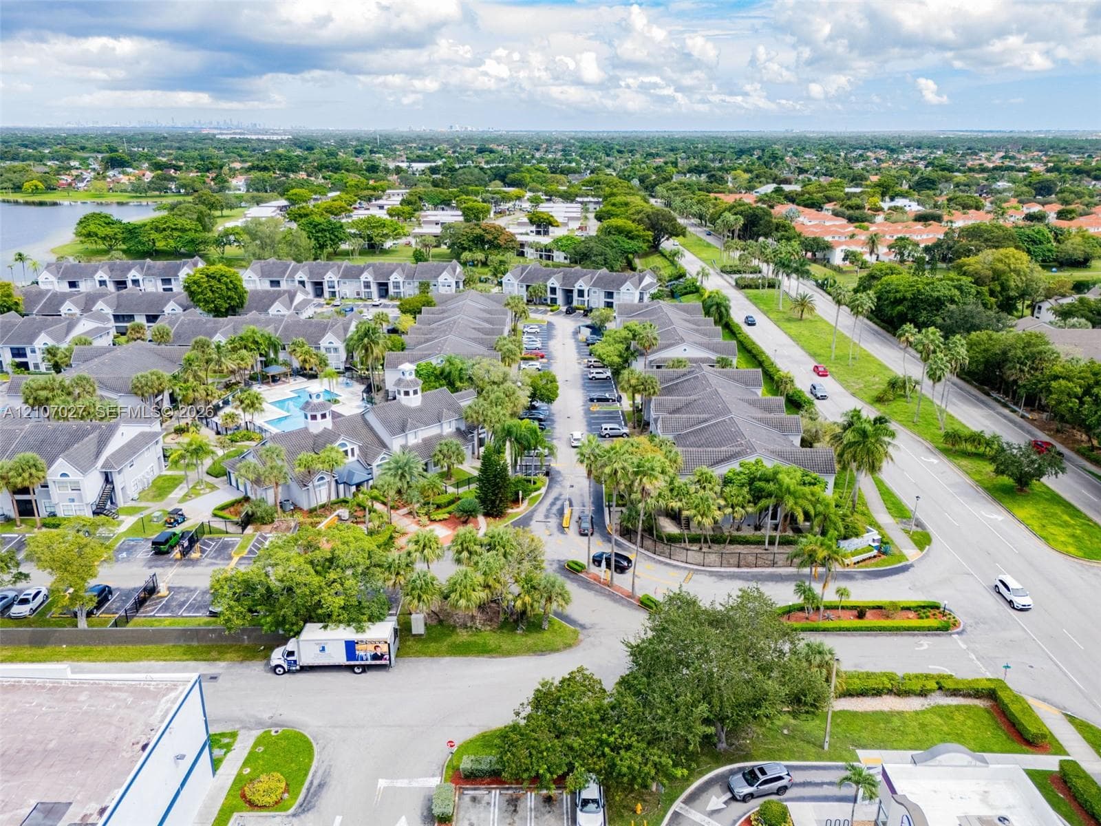 Aerial view of a residential community featuring low-rise buildings with grey shingle roofing, mature palm trees, landscaped green spaces, and a shared swimming pool
