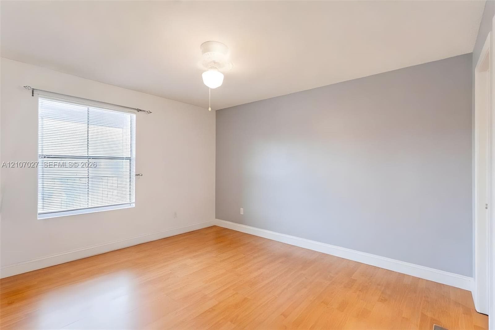 Neutral-toned room featuring wood-finish flooring, a single-pane window with horizontal blinds, and a central ceiling light fixture