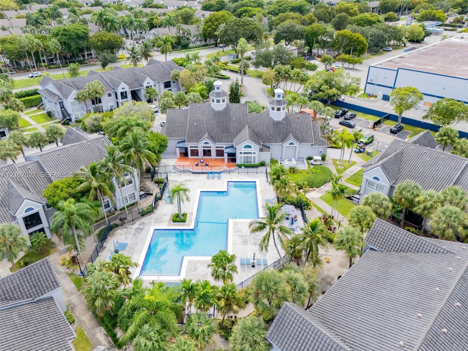 Aerial perspective of community grounds featuring an expansive swimming pool with hardscaped deck, multi-building architectural design with shingled roofing, and established palm tree landscaping