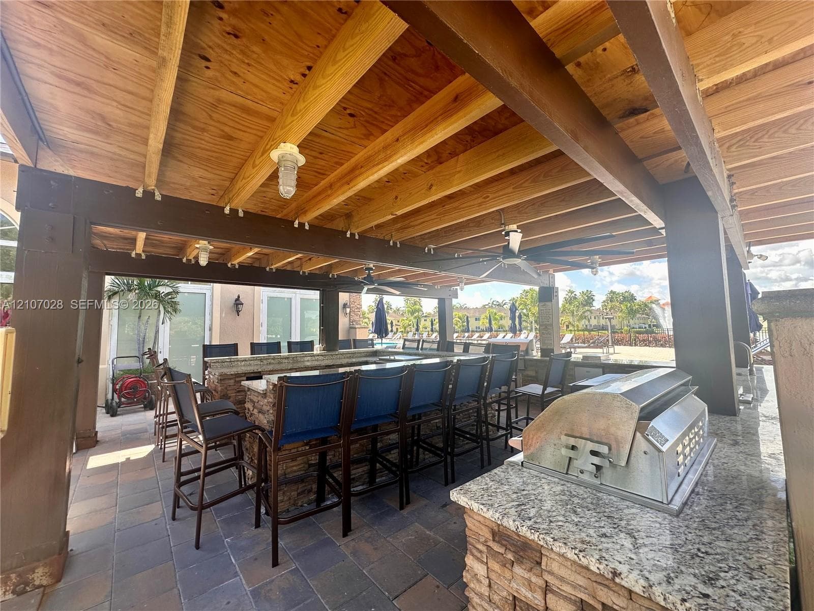 Covered outdoor kitchen featuring a stone-clad bar, granite countertops, built-in stainless steel grill, exposed wood-beam ceiling, and stone paver flooring