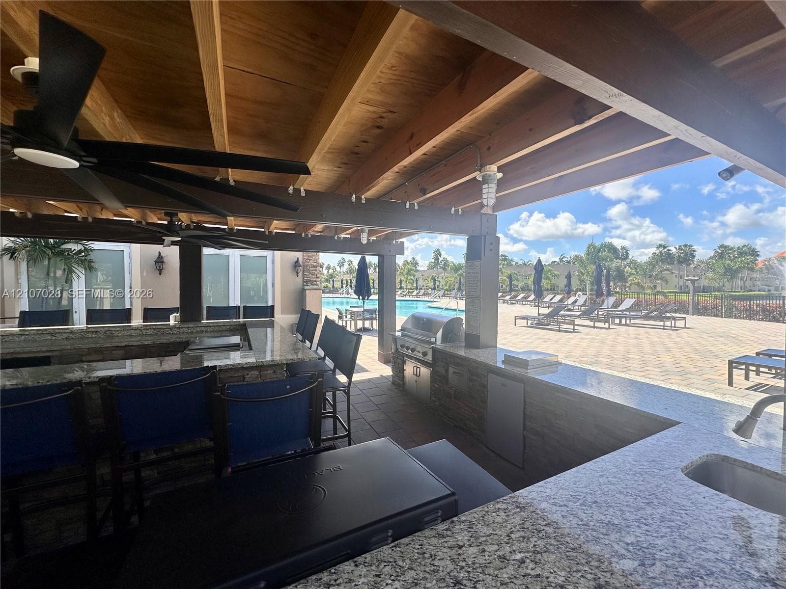 Covered outdoor kitchen featuring a stone-faced bar, granite countertops, built-in stainless steel grill, and exposed timber ceiling beams