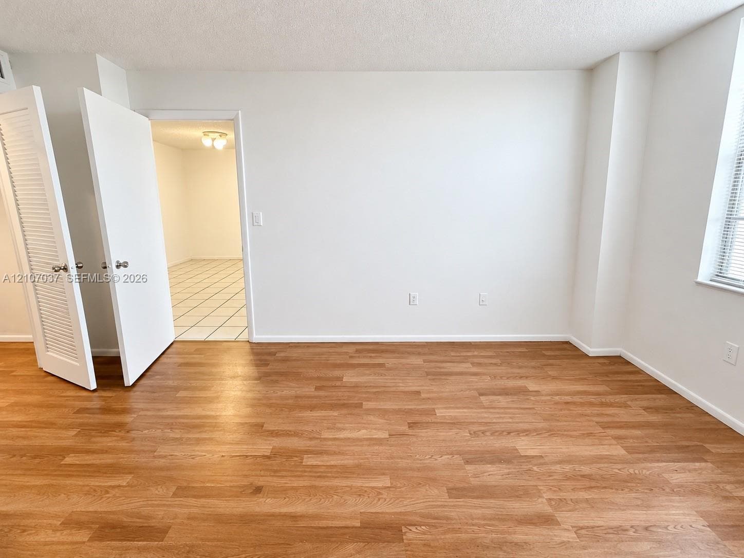 Interior room featuring wood-finish flooring, white walls, louvered closet doors, and large windows with horizontal blinds
