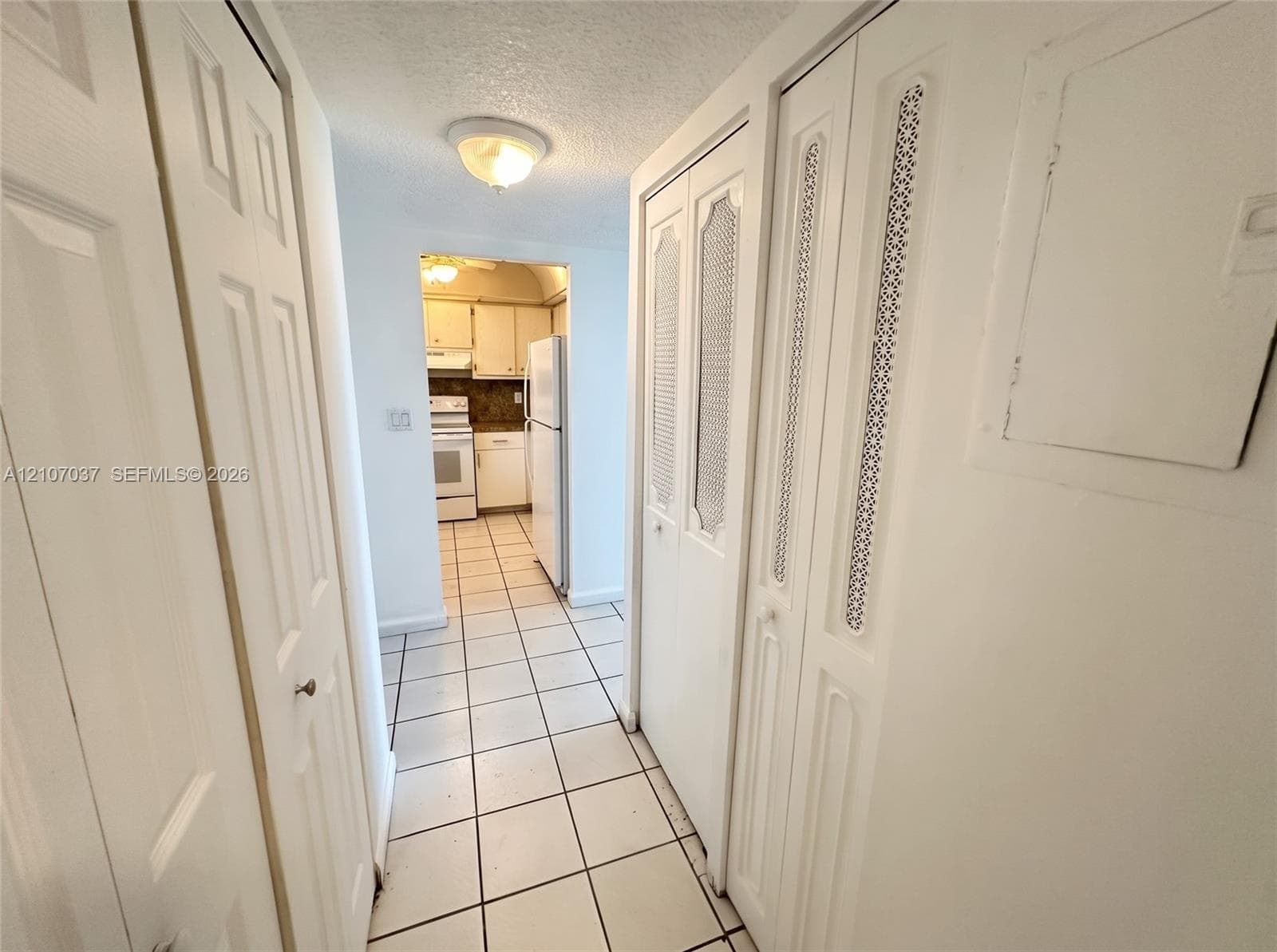 Hallway featuring dual sets of floor-to-ceiling bi-fold closet doors with decorative metal mesh paneling, white ceramic tile flooring, and a flush-mount ceiling fixture