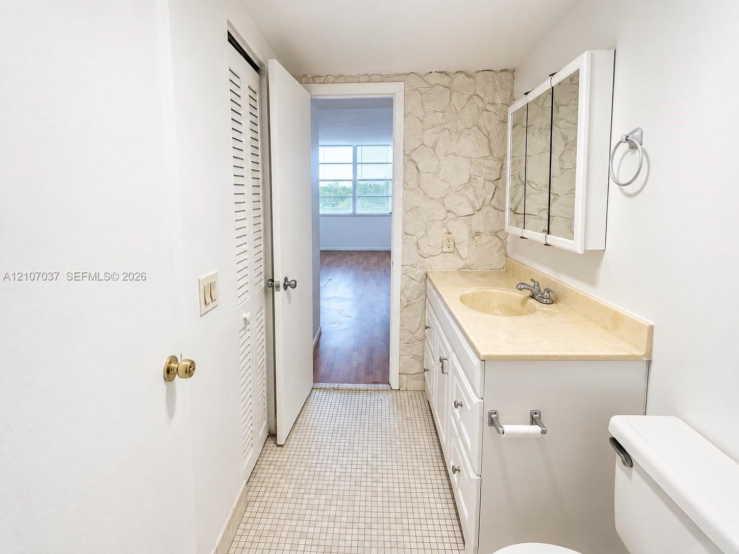 Bathroom featuring a textured stone accent wall, white vanity cabinet with a solid surface countertop, integrated oval sink, tri-fold mirrored medicine cabinet, and tiled flooring