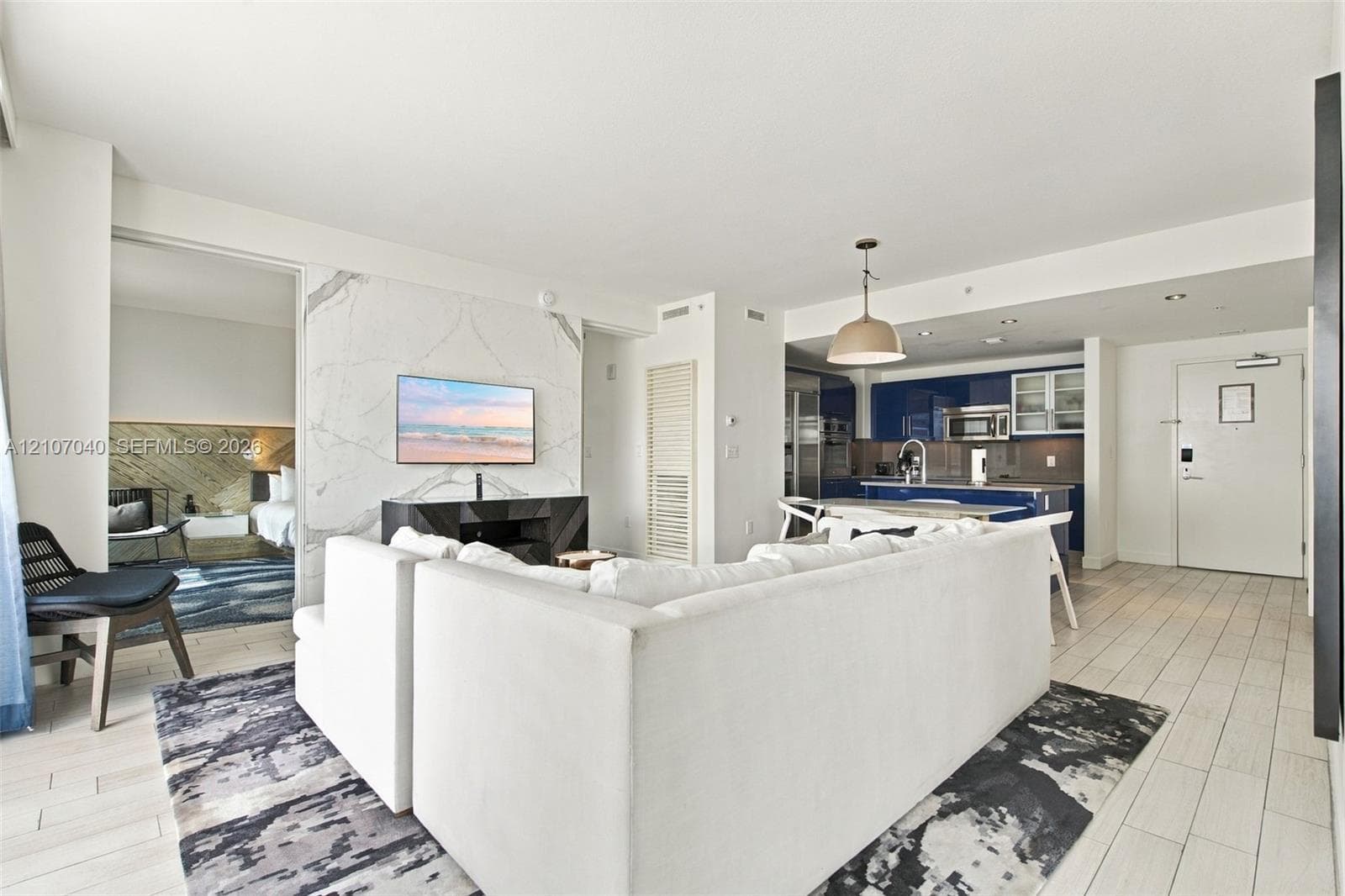 Open-concept living space featuring large-format tile flooring, a floor-to-ceiling stone-surround wall, glossy navy cabinetry, integrated kitchen appliances, and a polished metal pendant light fixture
