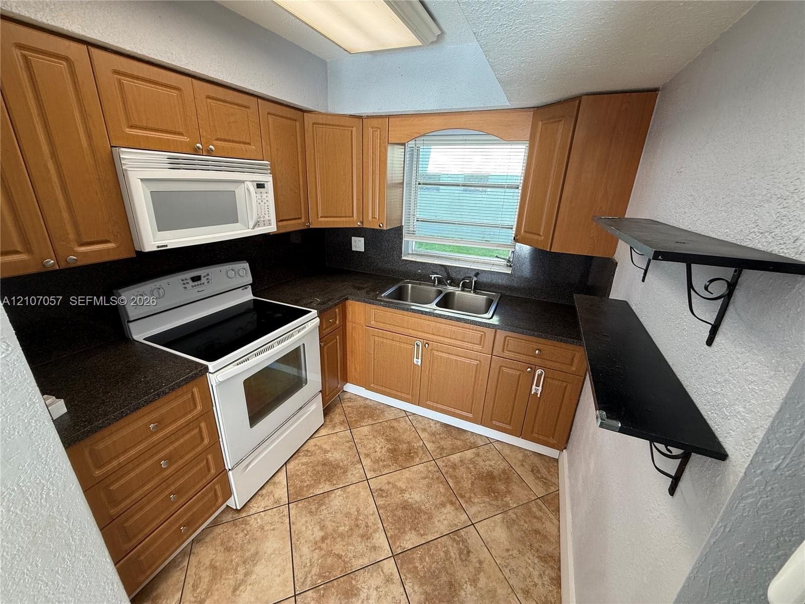 Kitchen featuring wood cabinetry, dark stone-finish countertops, tile flooring, built-in white microwave and range, and stainless steel double-basin sink