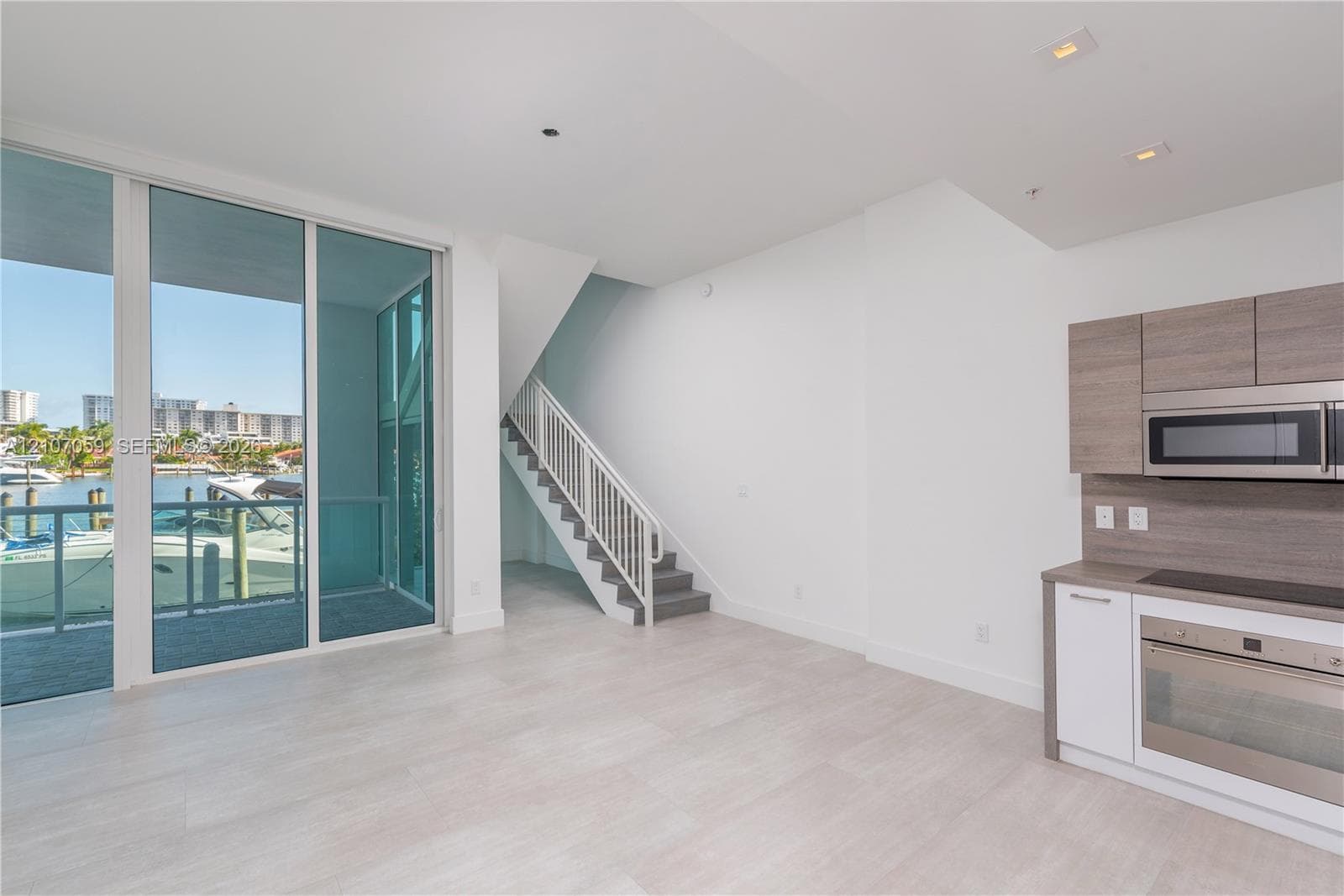 Floor-to-ceiling glass sliding doors, wood-finish cabinetry, built-in stainless steel appliances, recessed ceiling lighting, and a staircase with a metal railing