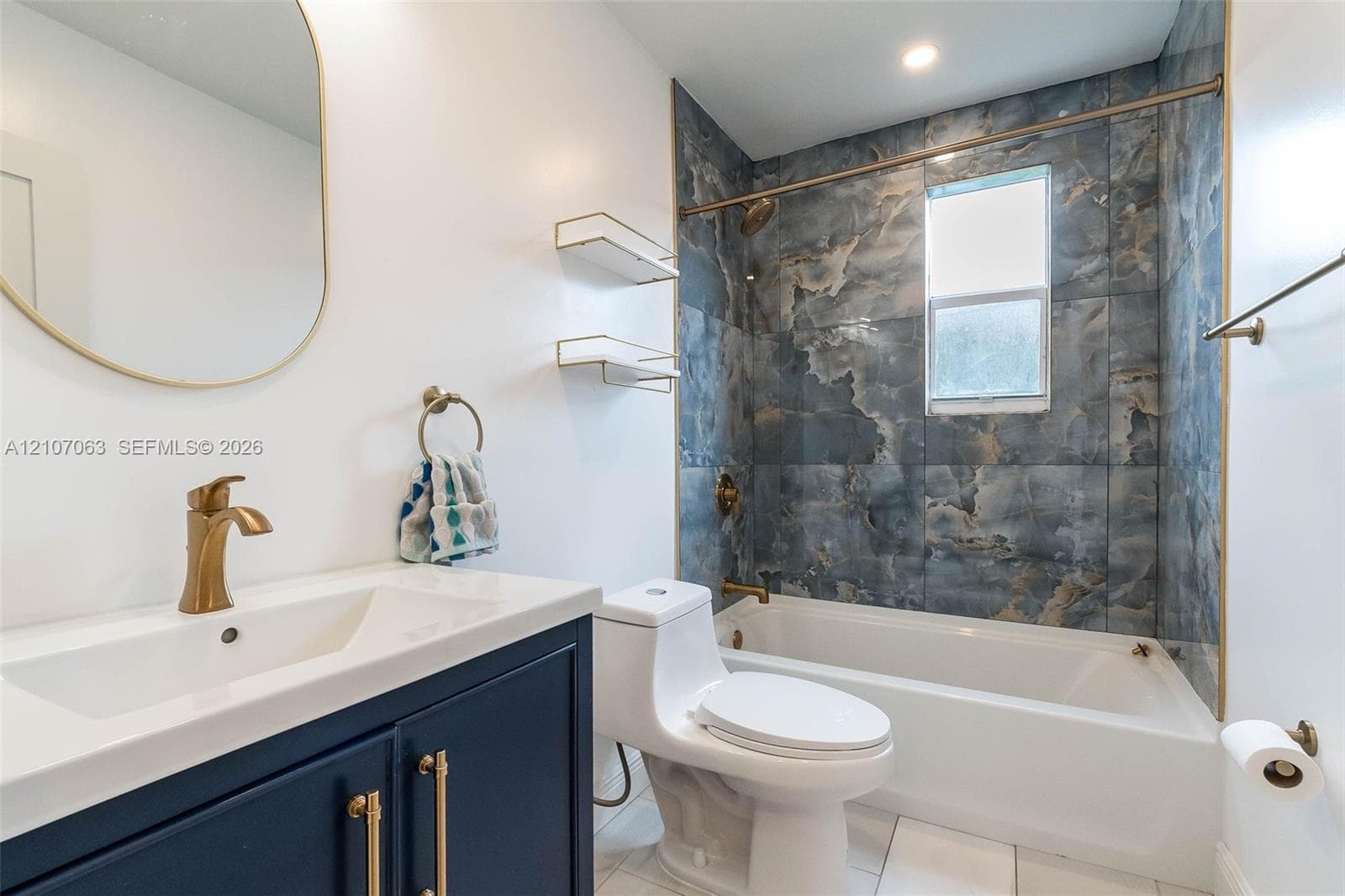 Bathroom featuring a navy blue vanity cabinet with brass hardware, an arched wall mirror, a white integrated sink, and a tub surround clad in blue-toned stone-patterned tile with brass fixture accents