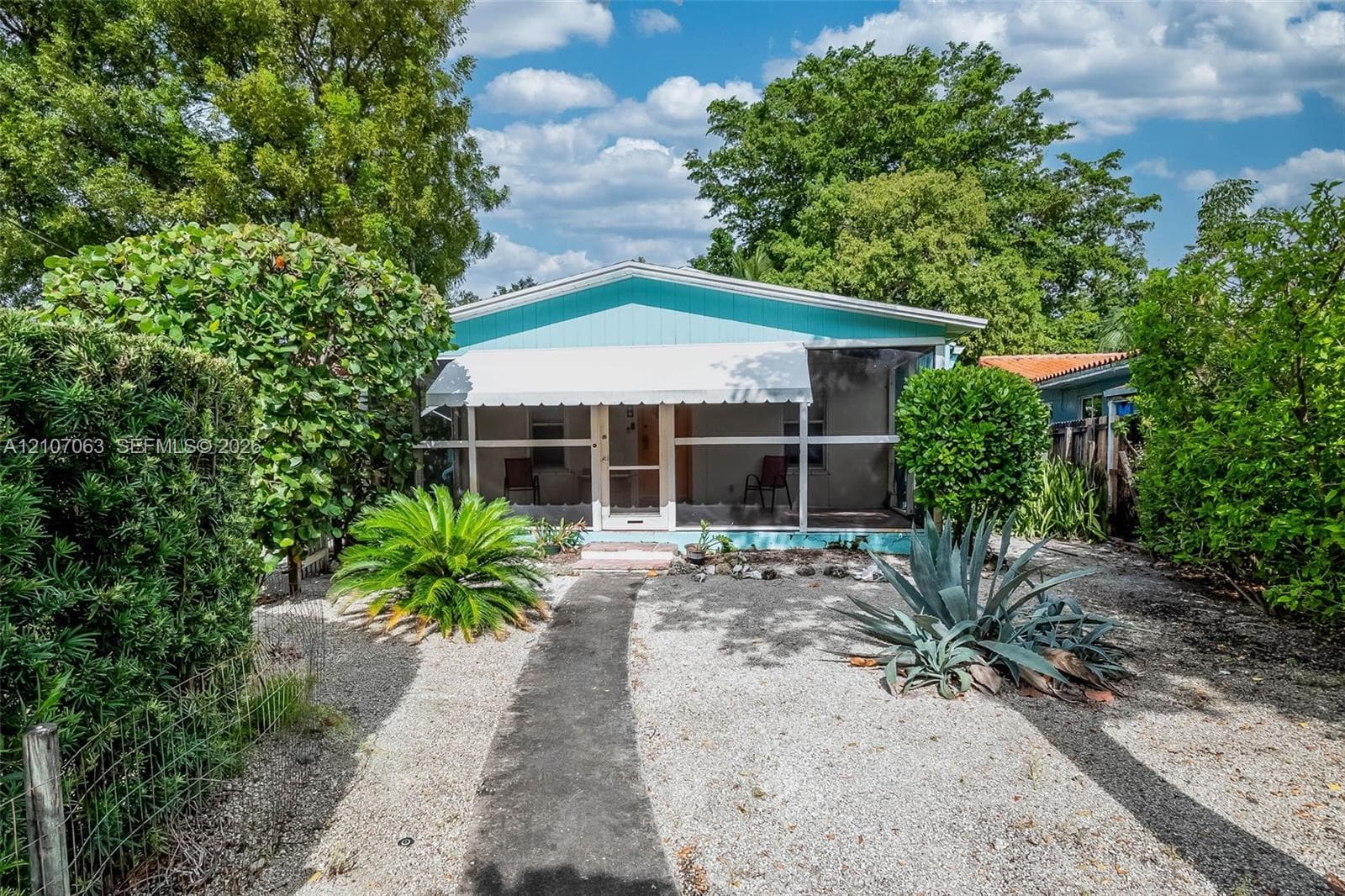 Exterior featuring a turquoise facade, covered screened porch, metal awning, and landscaped yard with decorative gravel and mature shrubbery