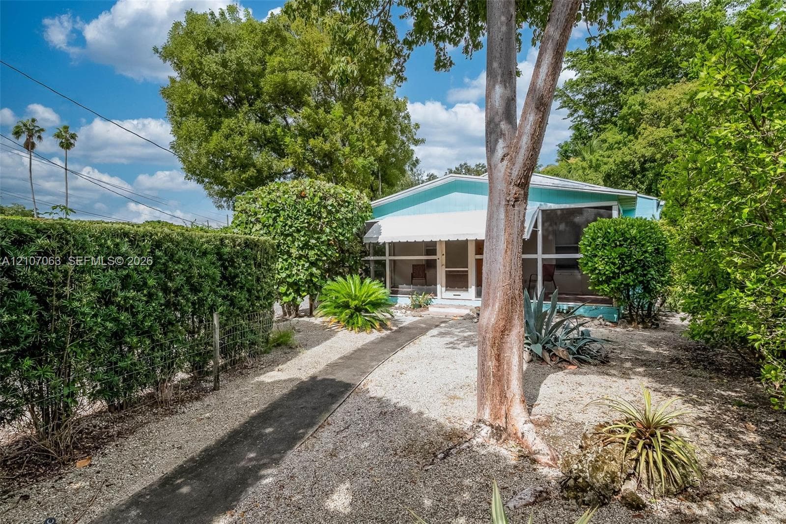 Exterior featuring a screened-in porch with metal awning, stone-covered front yard, paved walkway, mature trees, and privacy hedging