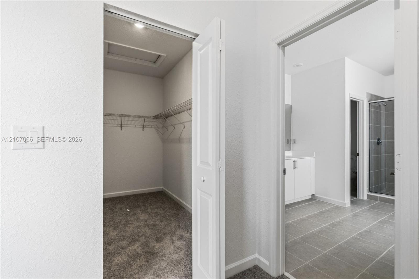 Walk-in closet with wire shelving and carpeting, featuring a bathroom with large-format tile flooring, white vanity cabinetry, and a glass-enclosed shower