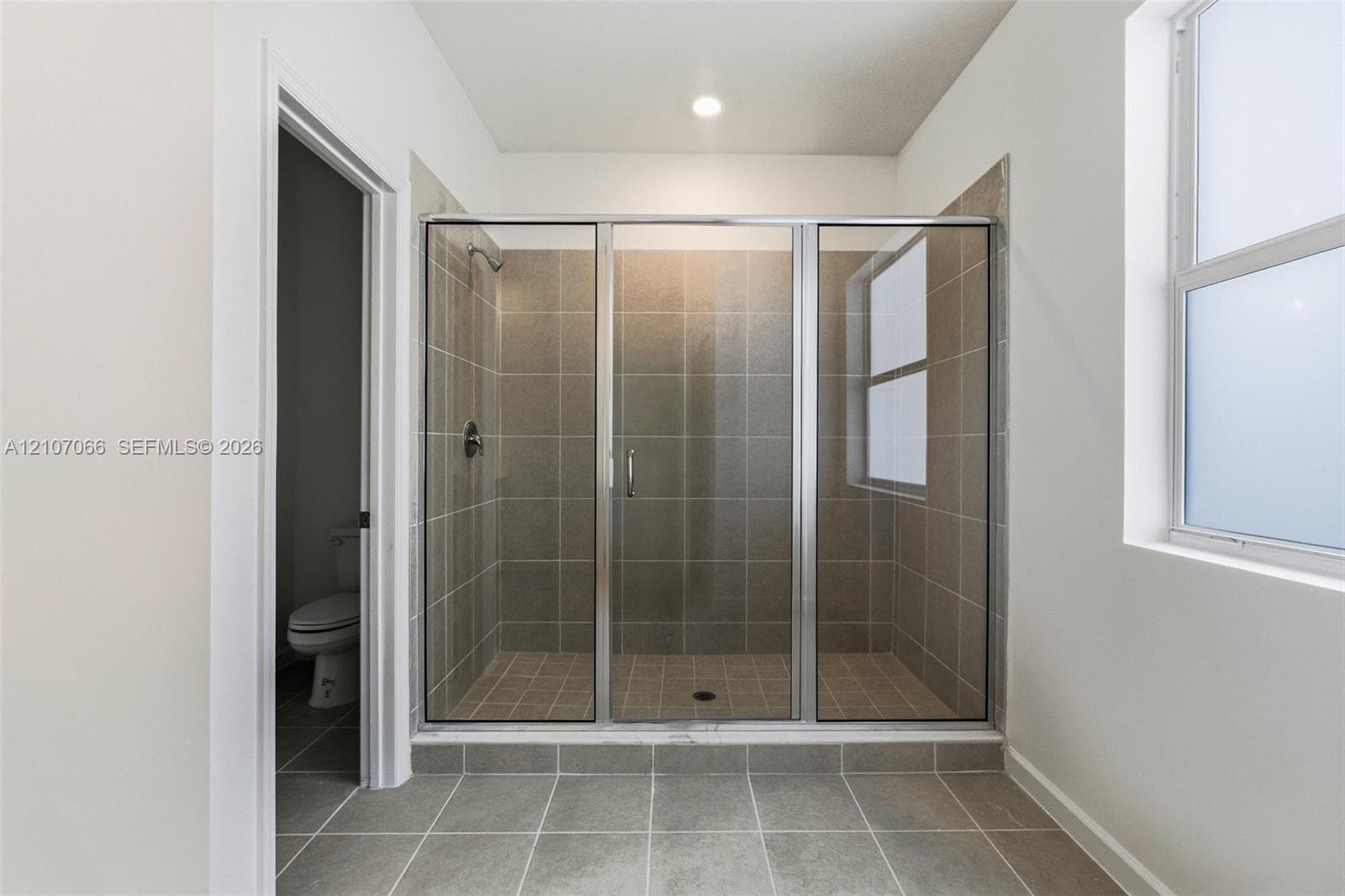 Walk-in shower featuring metallic framing, neutral ceramic wall and floor tiling, recessed lighting, and a window