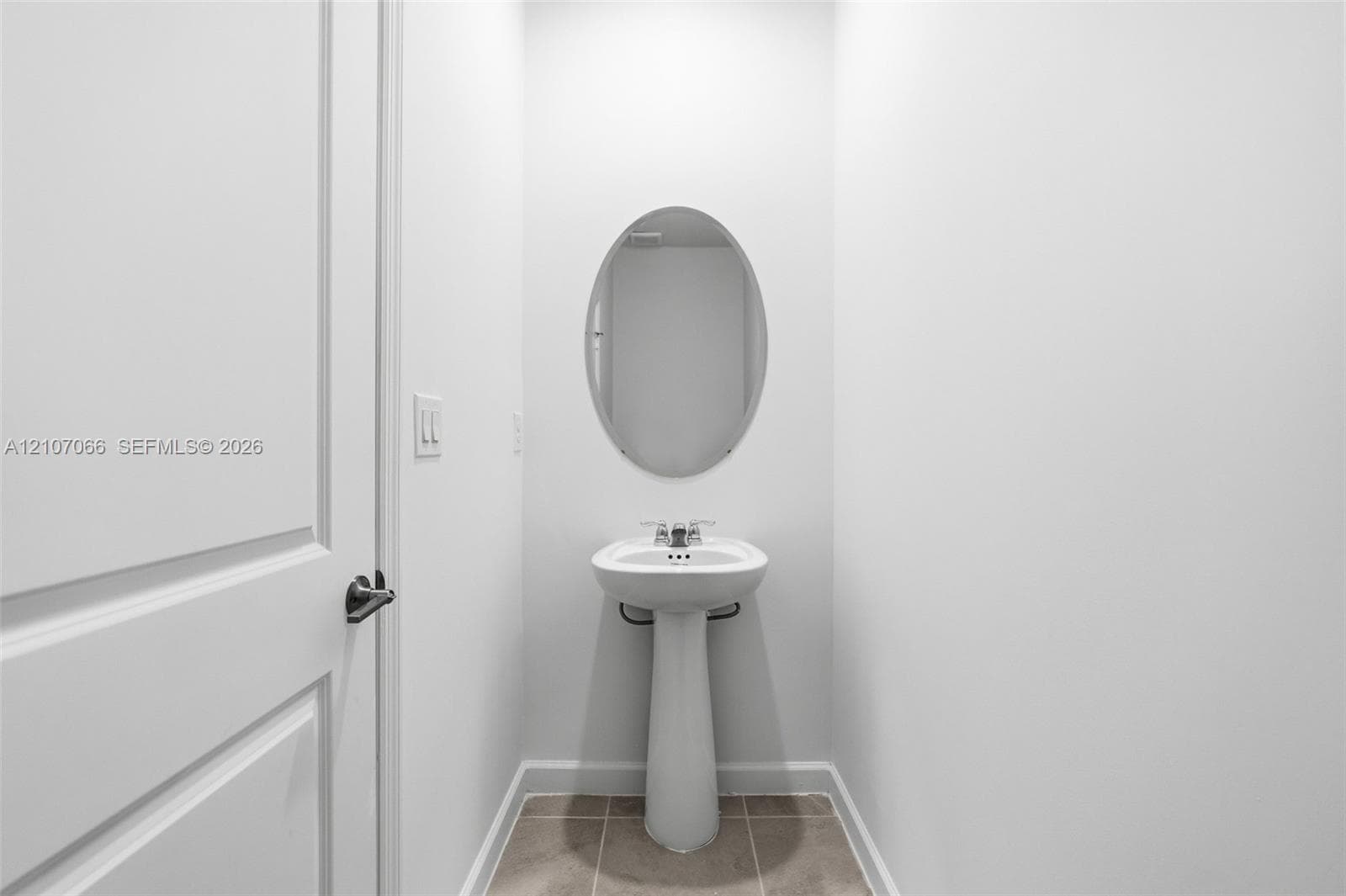 White pedestal sink with a dual-handle faucet, oval wall-mounted mirror, and tile flooring