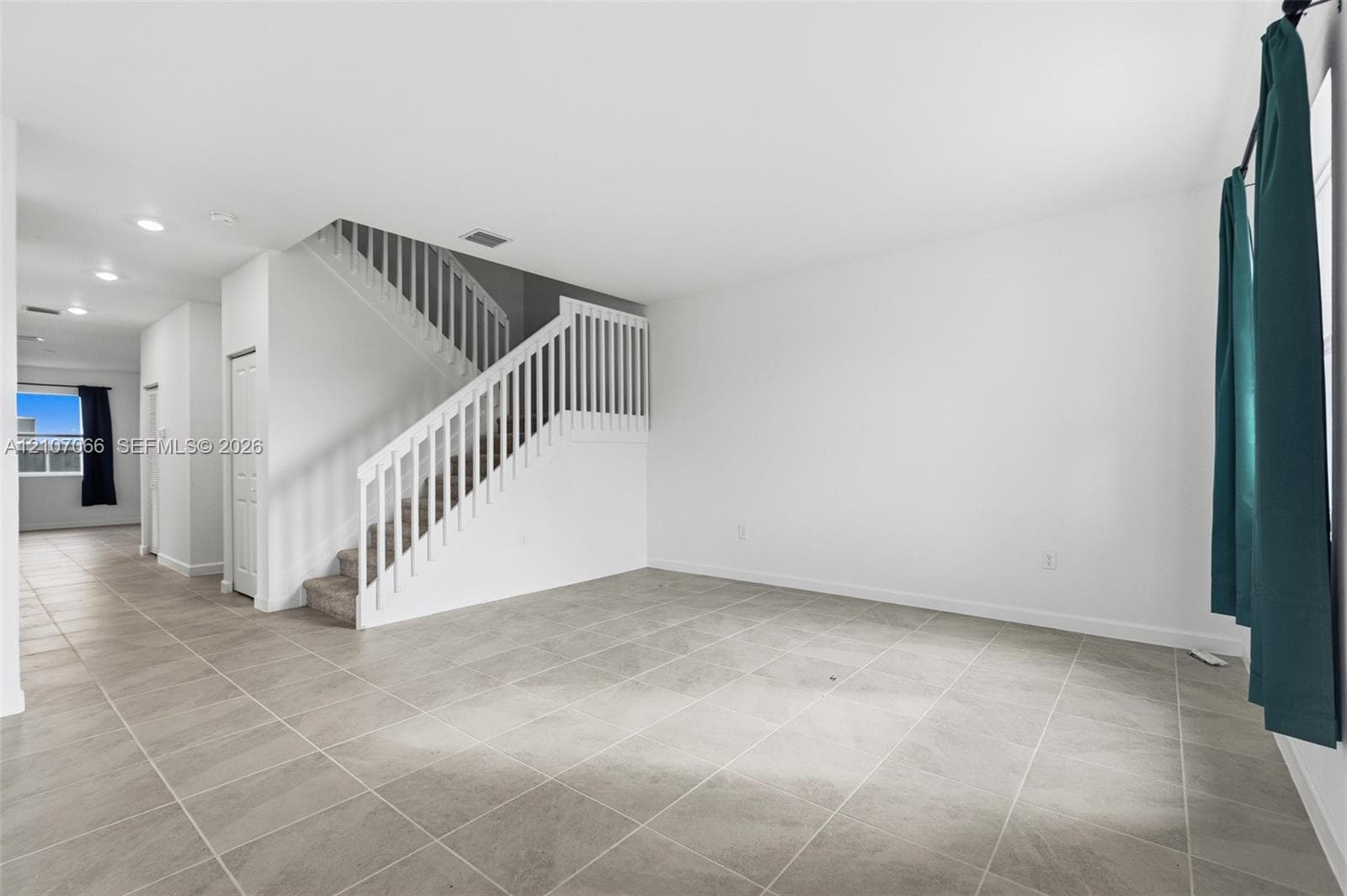 Large-format ceramic tile flooring, neutral wall palette, and white staircase featuring a wooden handrail and balusters