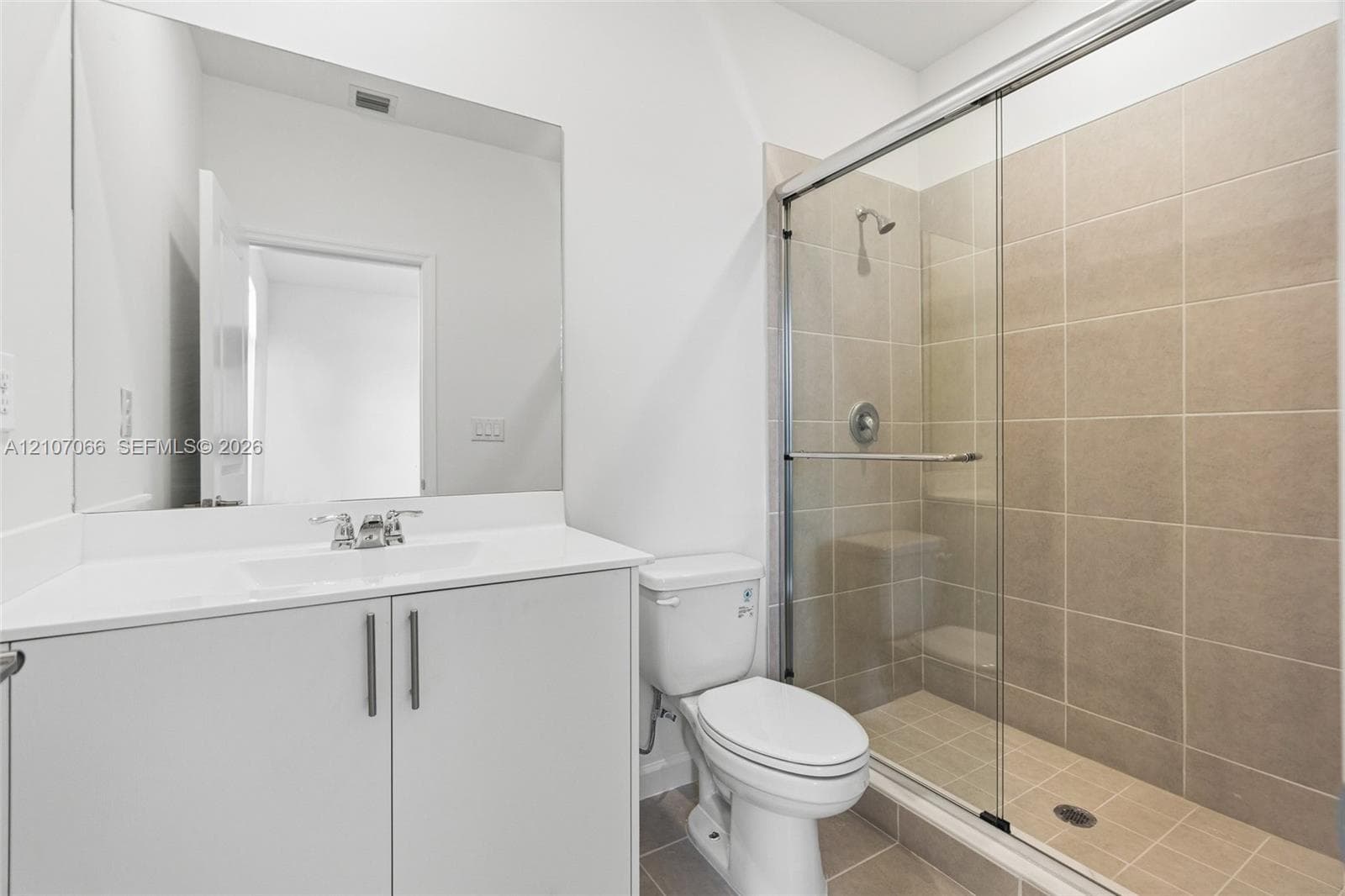 Bathroom featuring a white vanity cabinet with a solid surface countertop, an expansive wall-mounted mirror, a two-piece toilet, and a glass-enclosed shower stall with neutral-toned wall tile