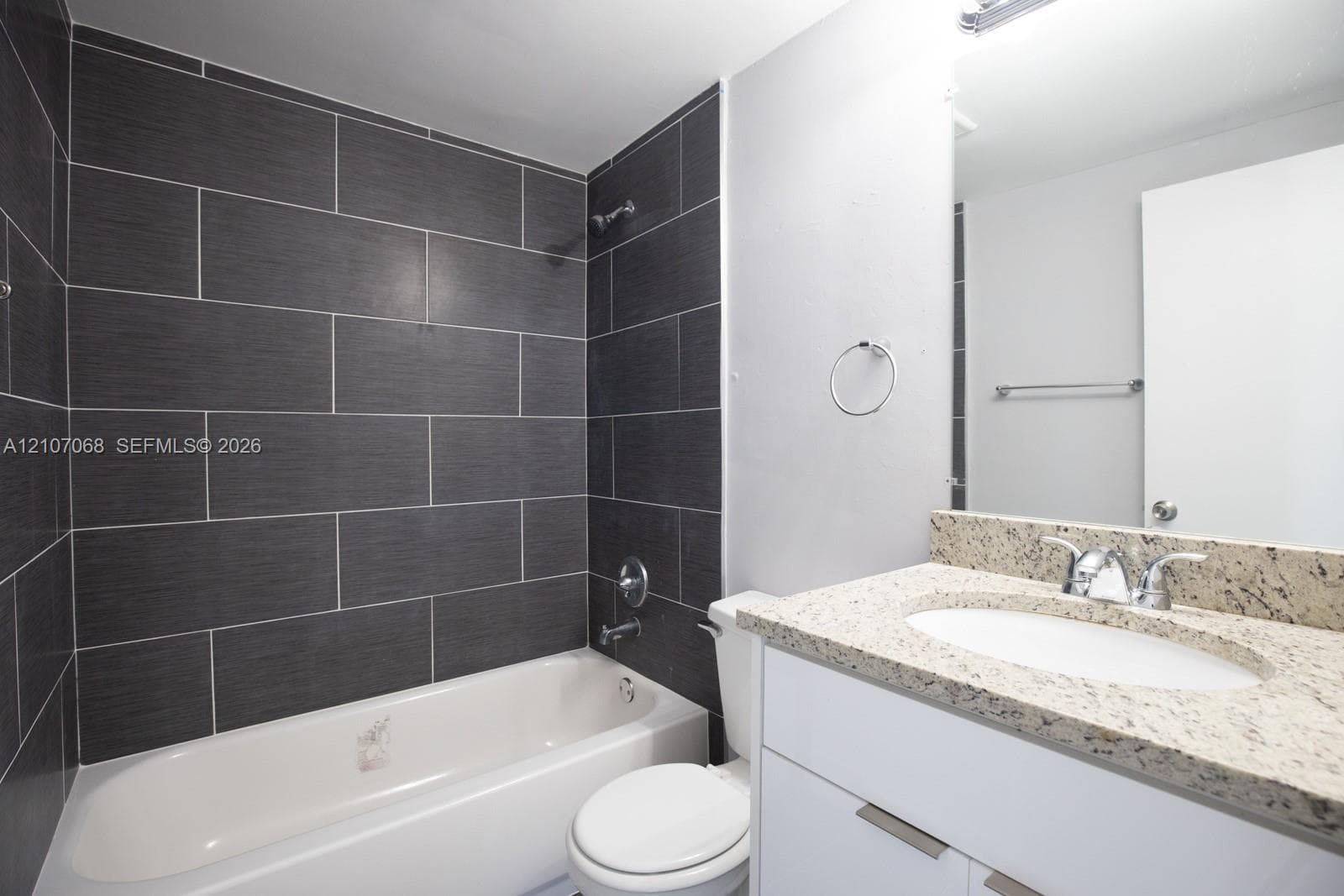 Bathroom featuring a tub-shower combination with dark large-format tile surround, white vanity cabinet, stone-finish countertop with undermount sink, and chrome fixtures