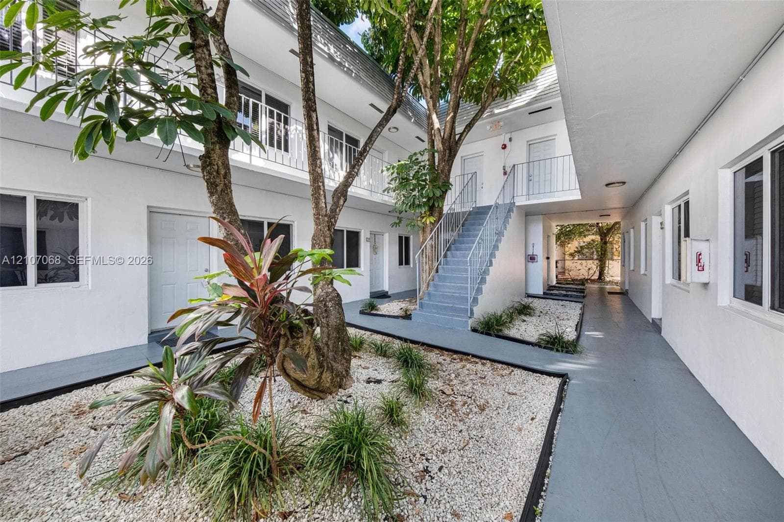 Courtyard featuring light-toned stucco exterior, metal staircase, balcony railings, mature trees, and rock-covered landscaping