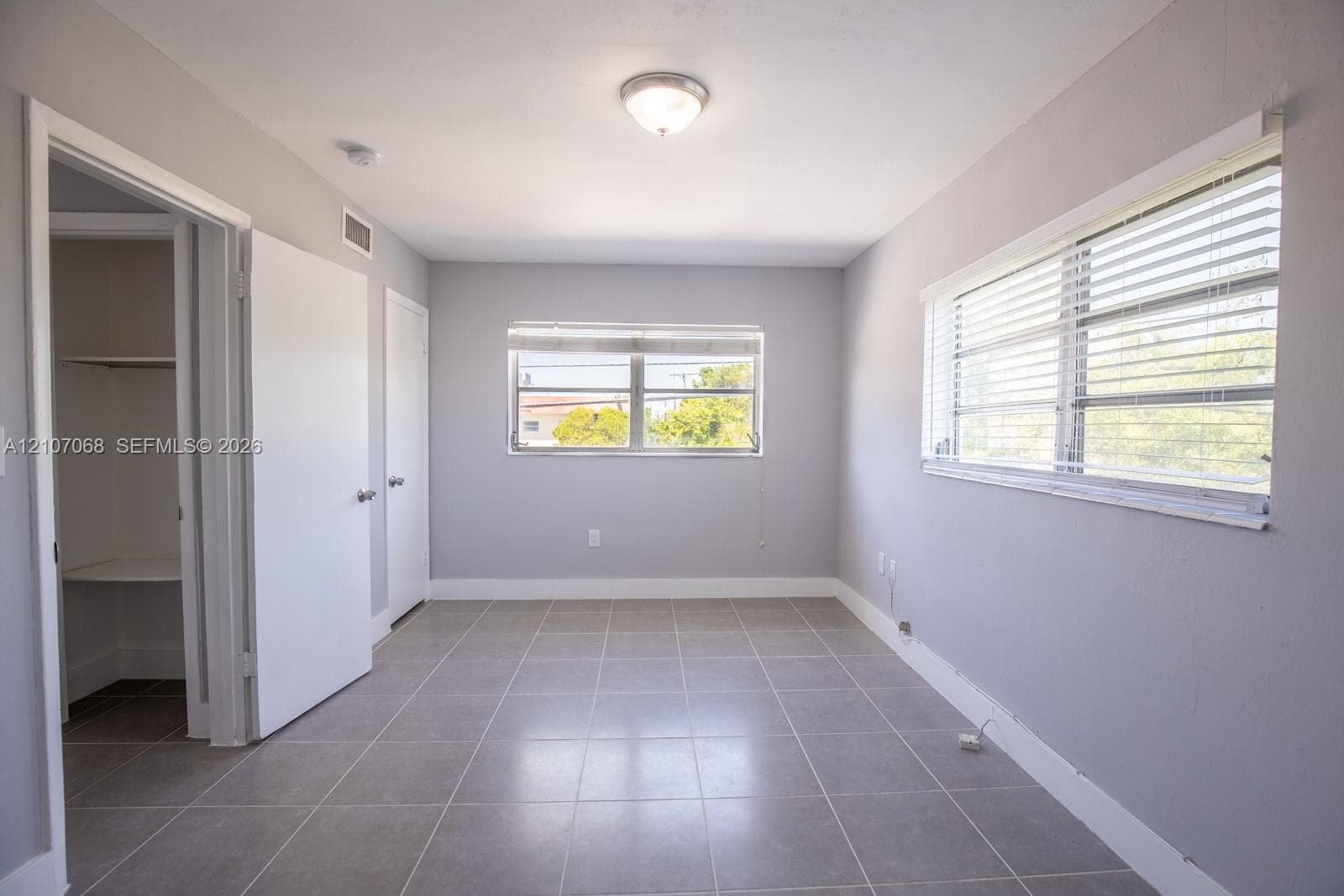 Neutral-toned room featuring large-format ceramic tile flooring, two windows with horizontal blinds, and a built-in closet with shelving