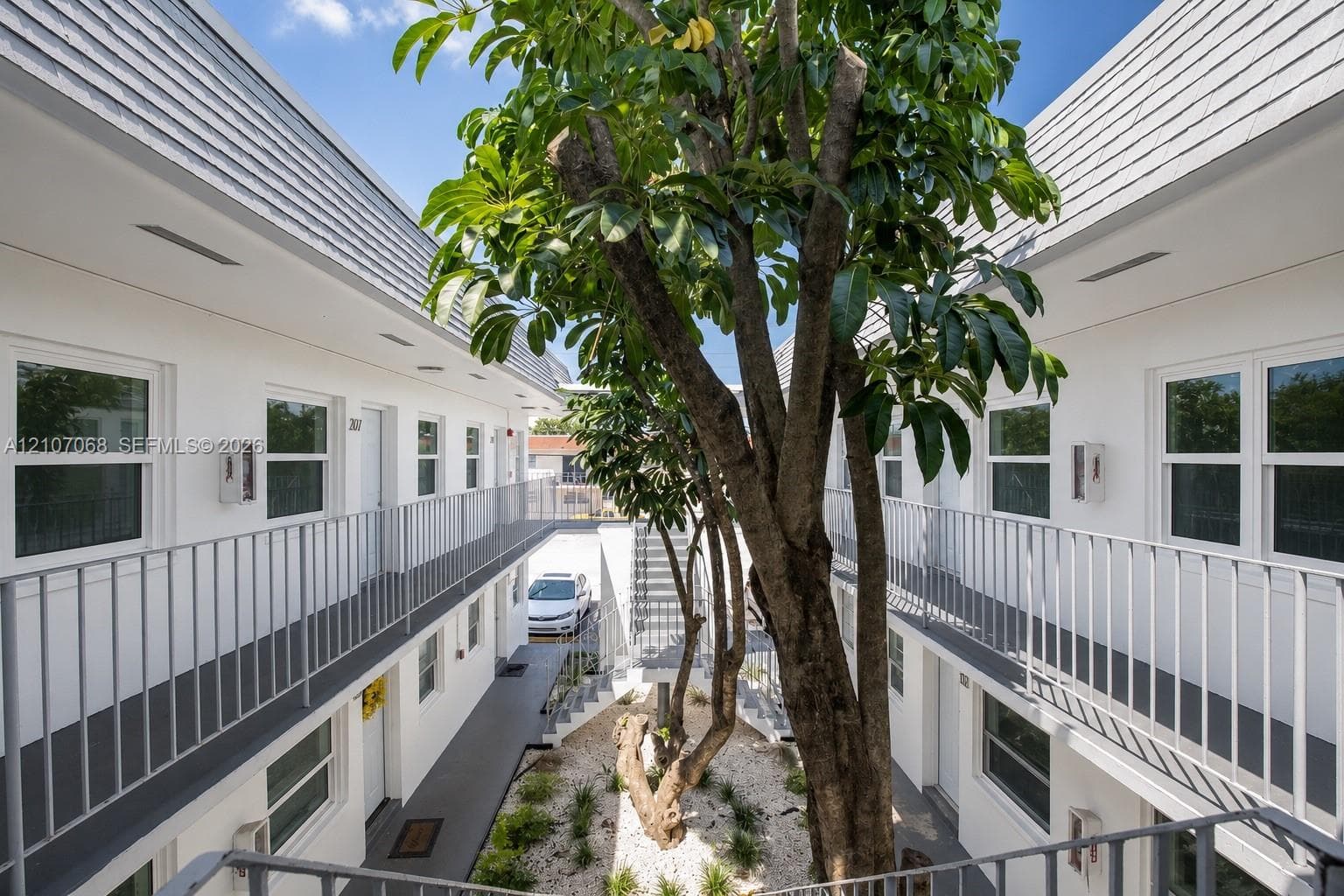 Exterior courtyard featuring white siding, metal railed walkways, and a central landscaped area with a mature tree and stone ground cover