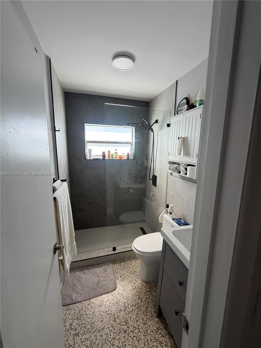 Bathroom featuring a glass-enclosed shower with a rainfall showerhead, large-format tile shower surround, terrazzo-style flooring, vanity cabinet with integrated sink, and white wall-mounted cabinetry