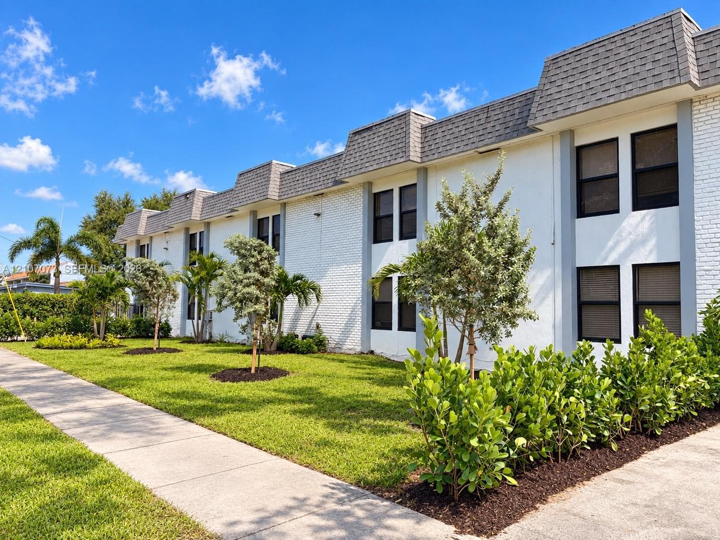 Two-story building exterior featuring white painted brick facade, mansard-style roofline, concrete pedestrian walkway, and mature landscaping
