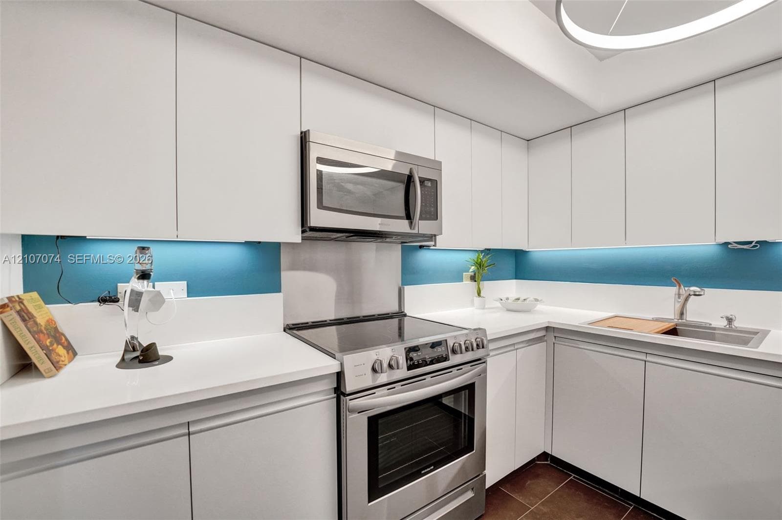 Kitchen featuring flat-panel white cabinetry, solid surface countertops, stainless steel appliances, integrated under-cabinet lighting, and a circular overhead light fixture