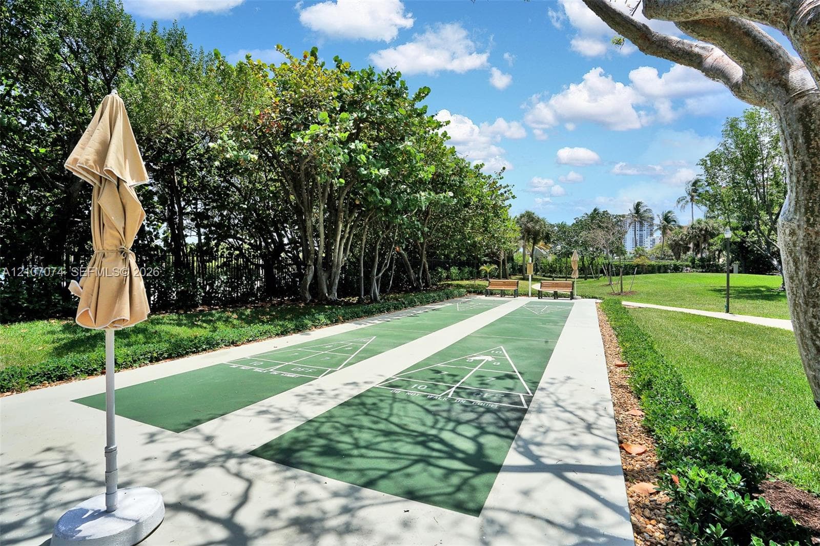 Dual shuffleboard court with painted green surfaces, concrete borders, landscaped perimeter, and mature shade trees