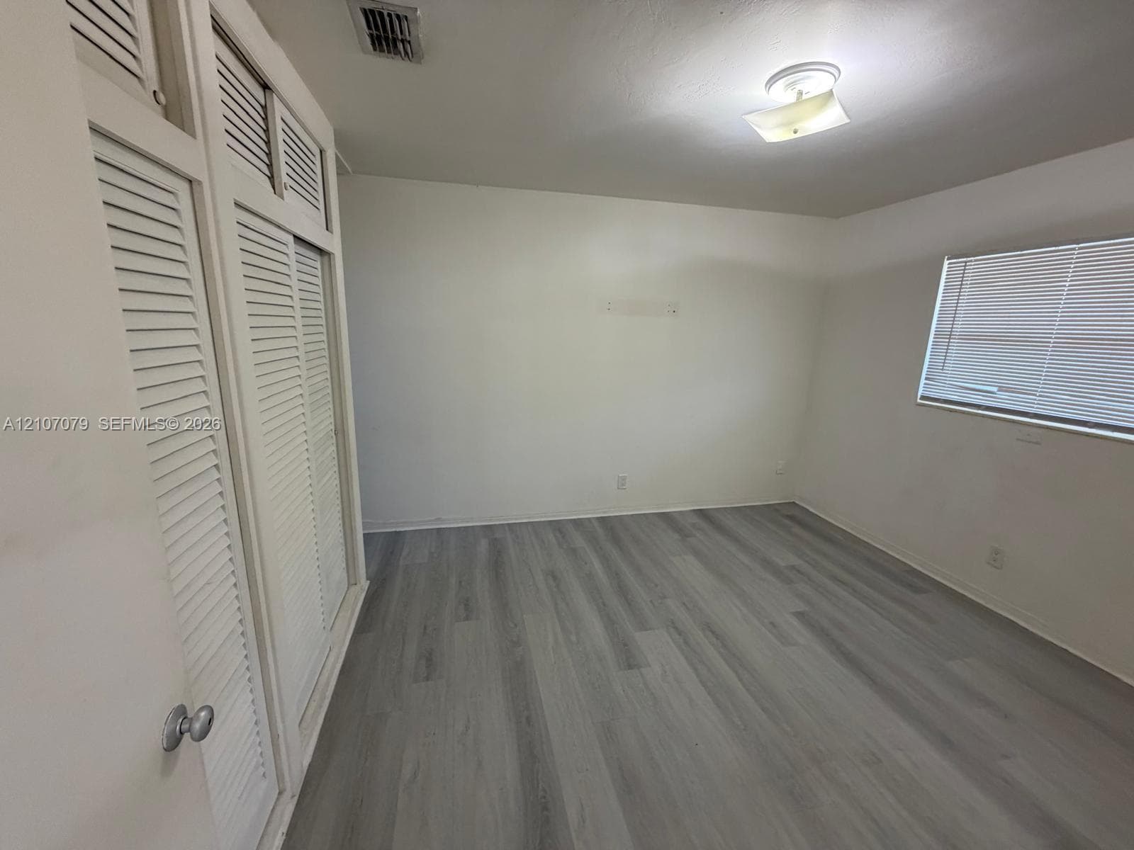 Room featuring wood-finish flooring, built-in louvered-door closets with overhead storage, a multi-pane window with horizontal blinds, and a centered flush-mount ceiling fixture