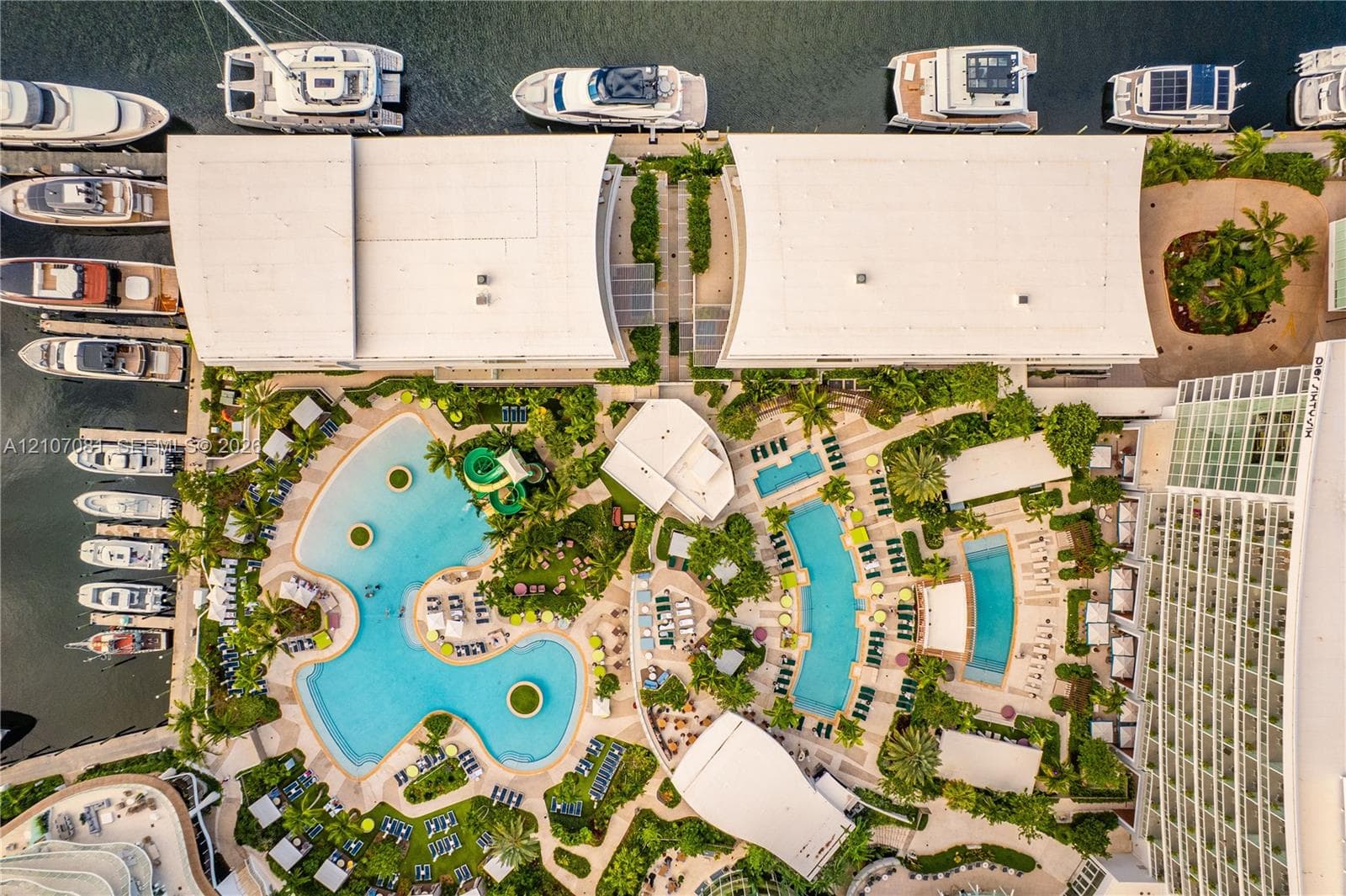 Aerial perspective of a waterfront property featuring multiple swimming pools, paved pool decks, lush landscaped grounds, and direct marina access