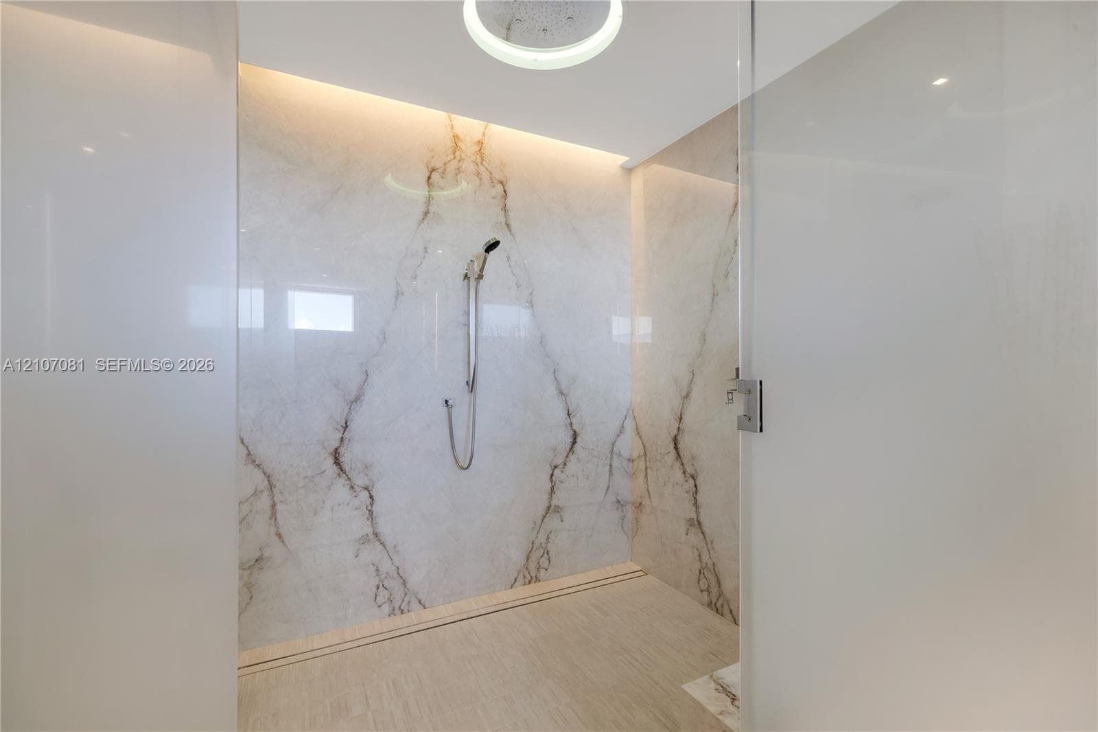Walk-in shower enclosure featuring large-format veined stone wall panels, integrated ceiling lighting, a handheld shower fixture, and a glass door