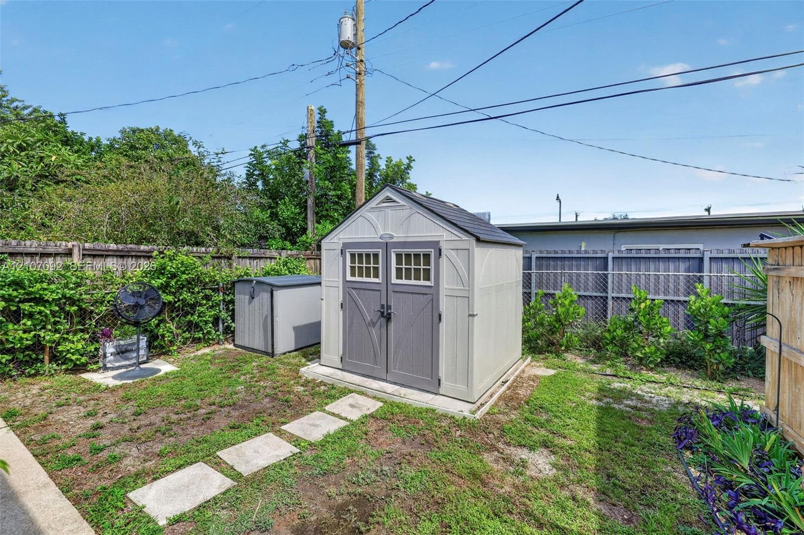 Exterior storage structure featuring a gable roof, double doors with glass paneling, and a reinforced foundation pad, situated within a fenced backyard