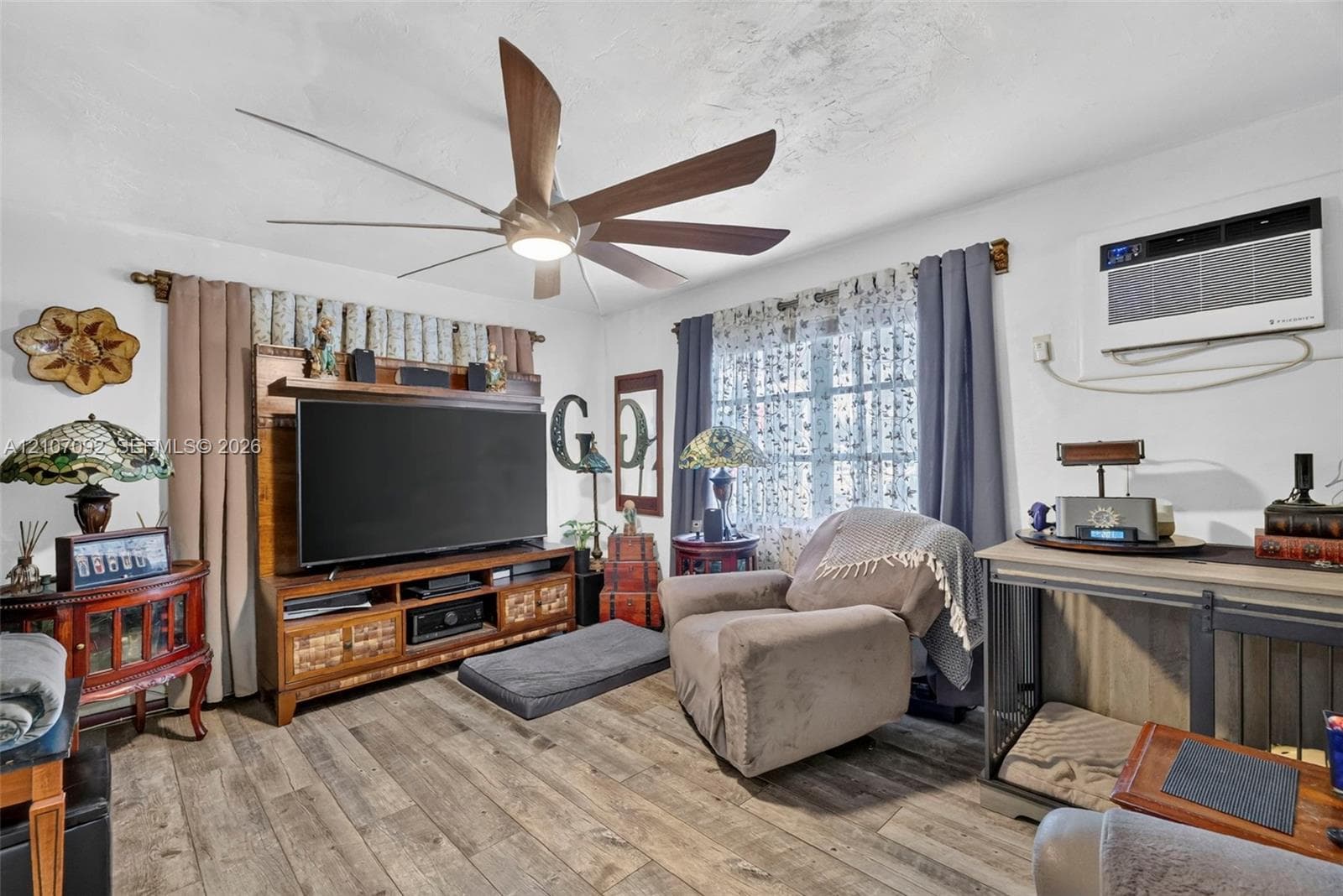 Living area featuring wood-finish flooring, a multi-blade ceiling fan with integrated lighting, and a wall-mounted air conditioning unit