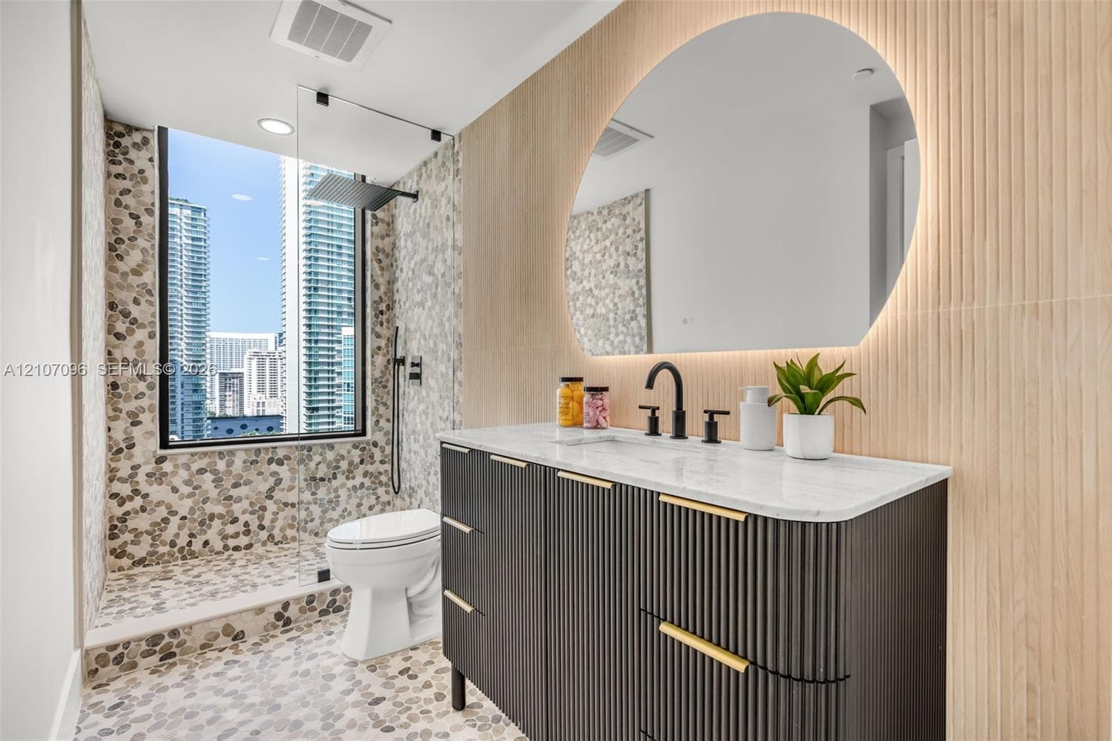 Custom dark fluted vanity with gold-tone hardware and light-toned stone countertop, integrated backlit arched mirror, floor-to-ceiling pebble tile shower surround, and matte black rain shower head