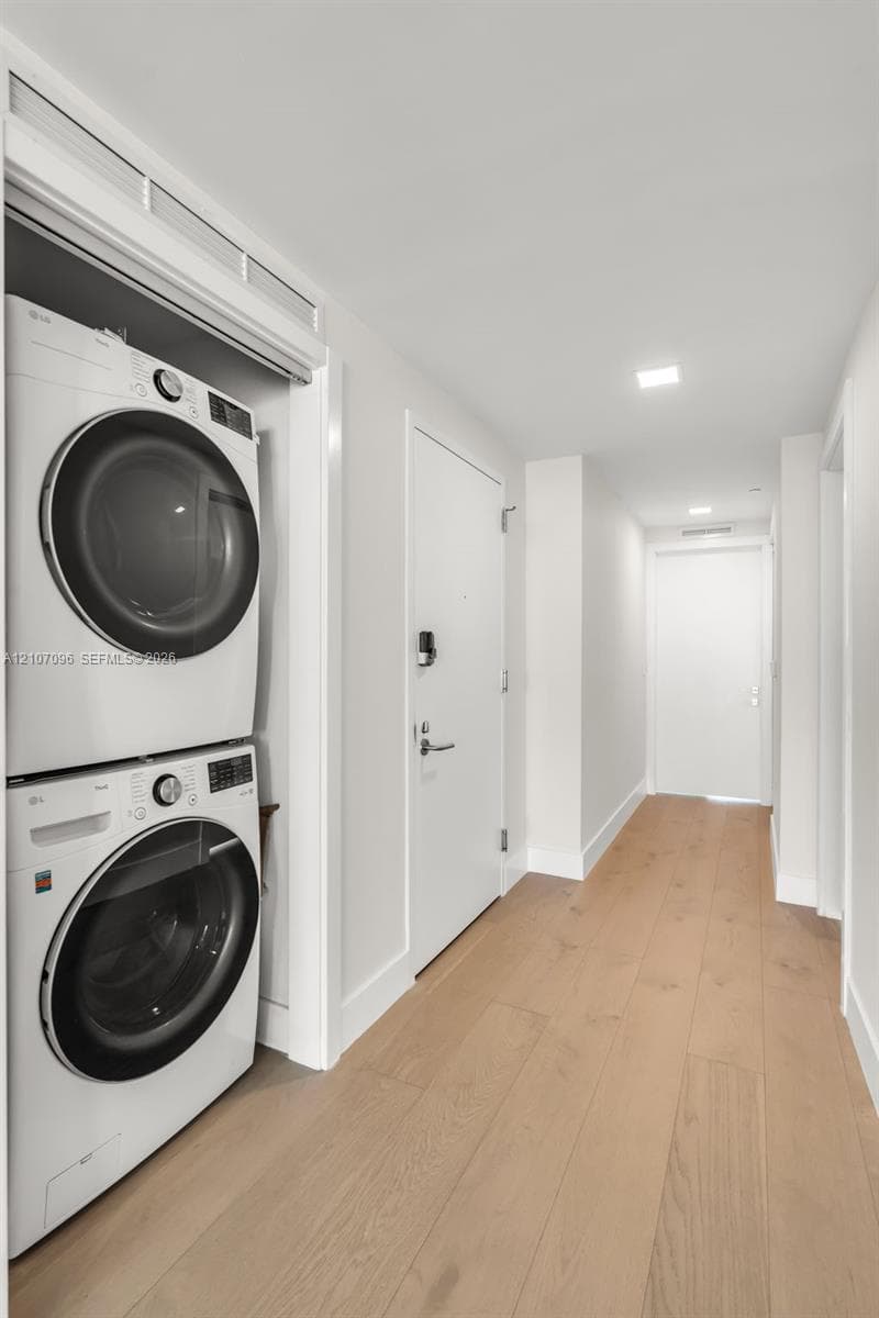 Stacked white front-load laundry appliances, light-toned wood-finish flooring, recessed ceiling lighting, and solid core doors