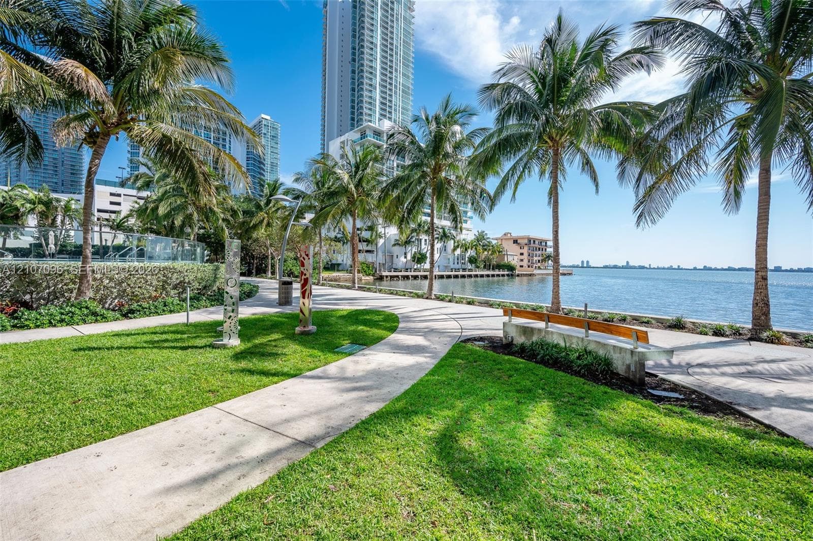 Waterfront grounds featuring a concrete walking path, manicured lawn, palm trees, and built-in bench seating
