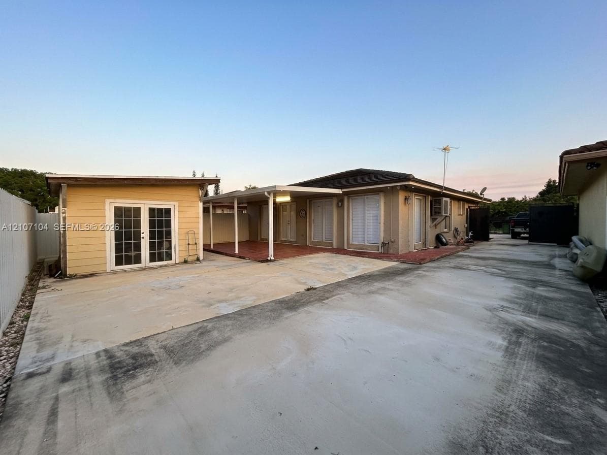 Concrete driveway, detached accessory structure with French doors, covered patio with tile flooring, and exterior wall-mounted lighting