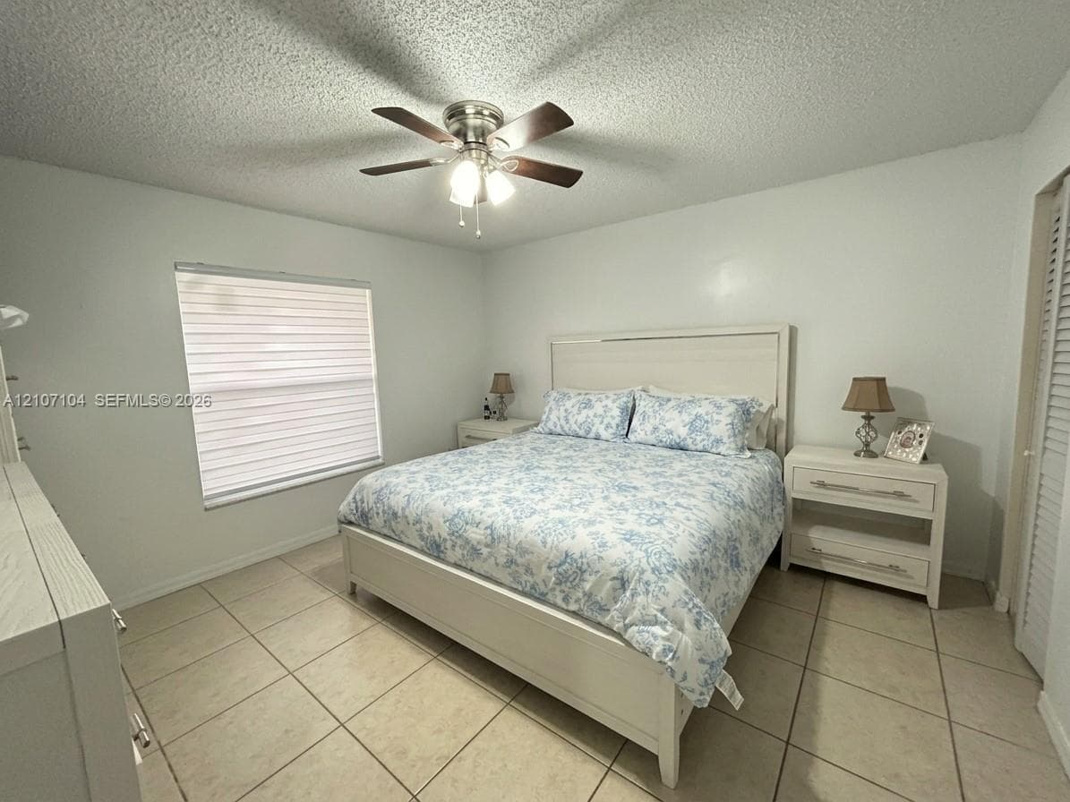 Neutral-toned ceramic tile flooring, ceiling fan with integrated light fixture, window with horizontal blinds, and louvered closet doors