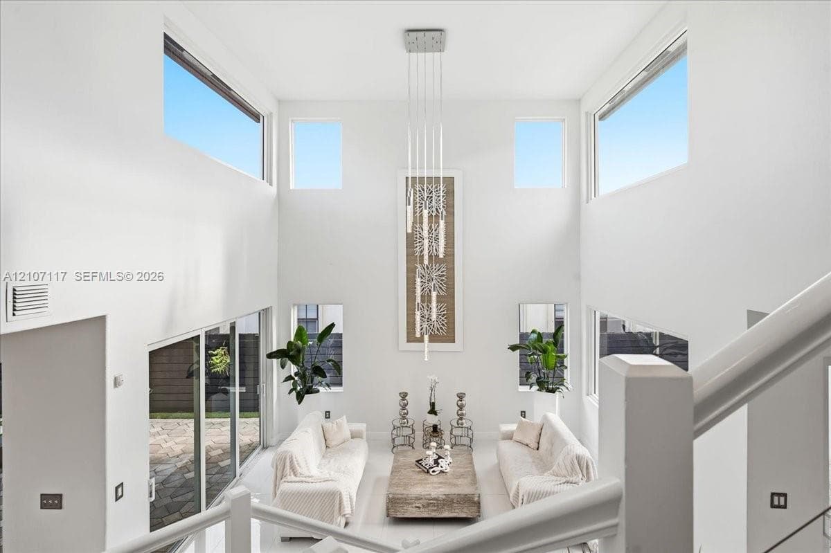 Double-height ceilings, expansive window configuration, multi-level glass slider access to exterior patio, and tiered contemporary pendant lighting