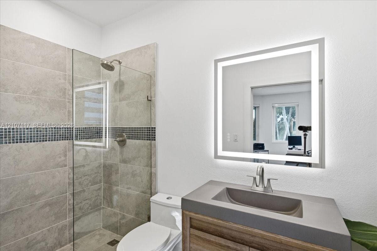 Walk-in shower with stone-finish tile and a mosaic tile inlay, wood-finish vanity with a molded sink, and a wall-mounted LED-backlit mirror